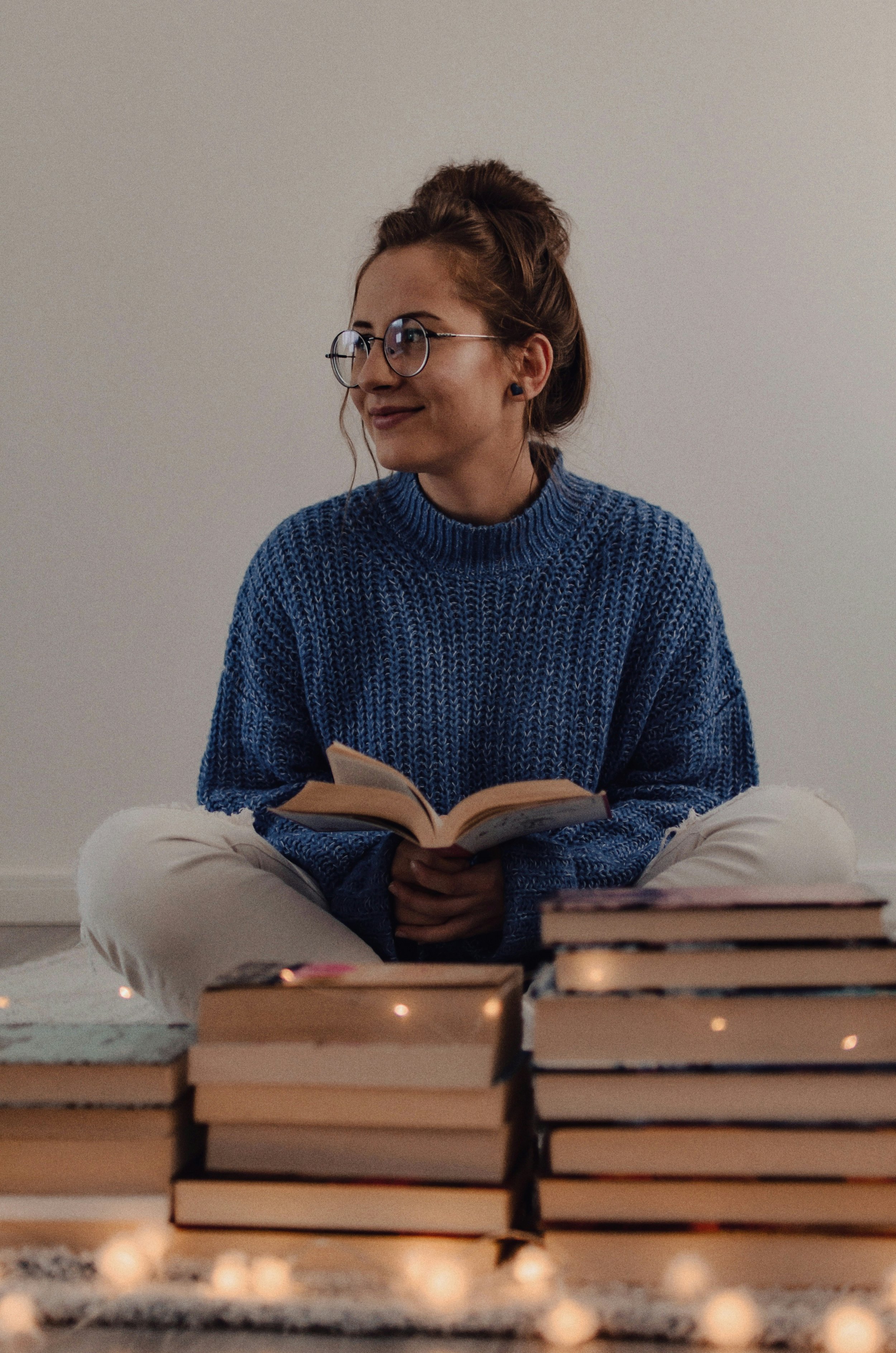 A woman sitting cross-legged on a carpeted floor, reading a book, dressed in a blue knit sweater and beige pants, with several stacked books in front of her.