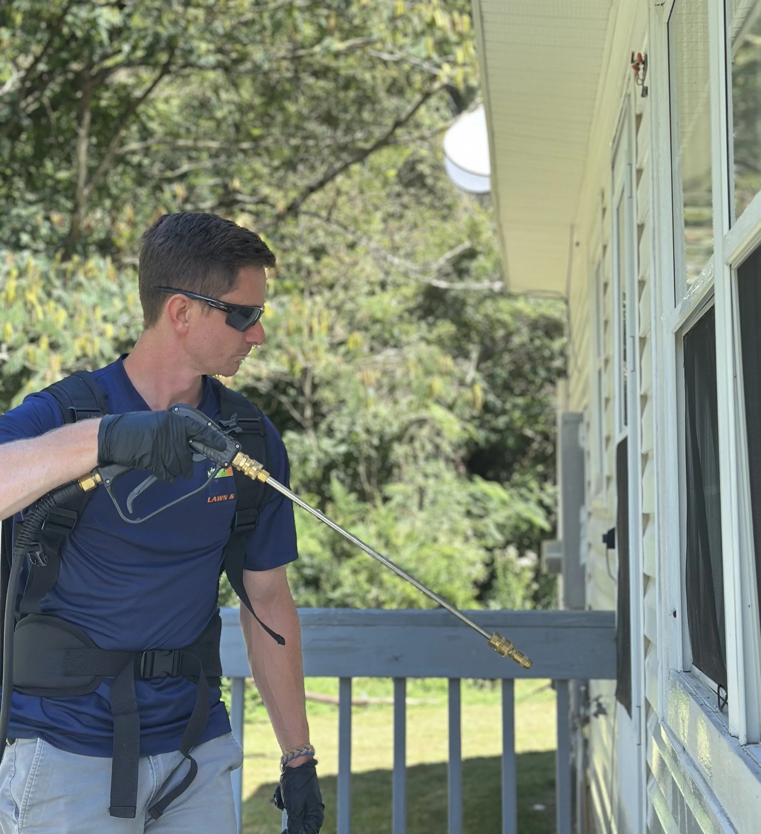 A man wearing sunglasses, gloves, a backpack, and a blue shirt is using a spray wand to clean the exterior of a house near a window on a sunny day.