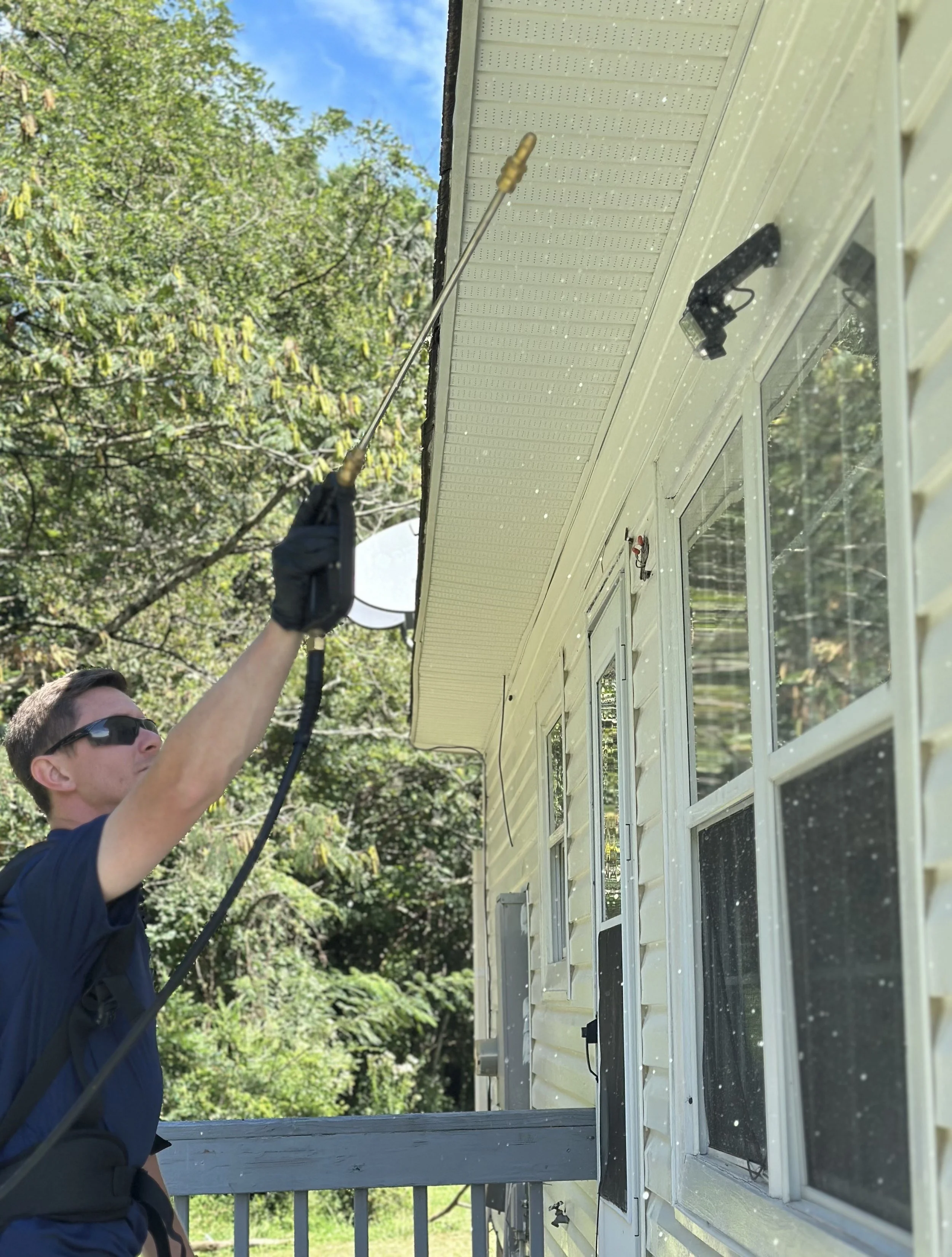 A man wearing sunglasses and a black glove is pressure washing the exterior of a house.