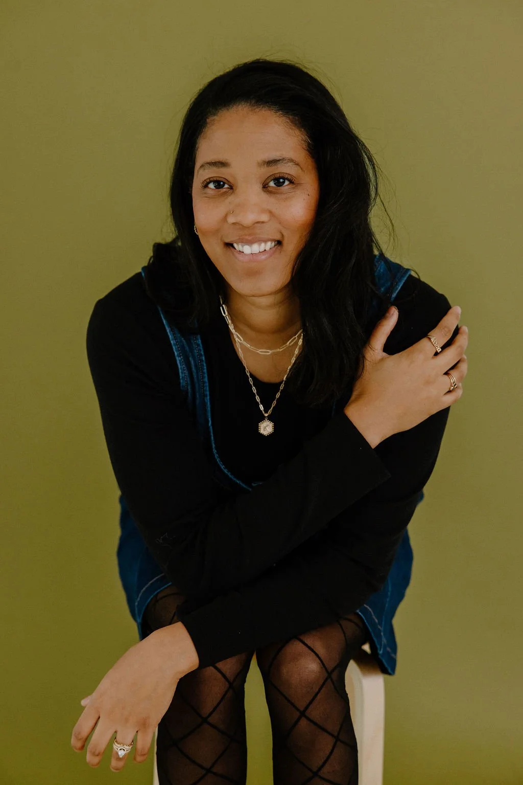 A woman with long black hair, smiling, sitting on a stool against a green background, wearing a black top with blue denim overalls, fishnet stockings, and layered necklaces, with rings on her fingers.