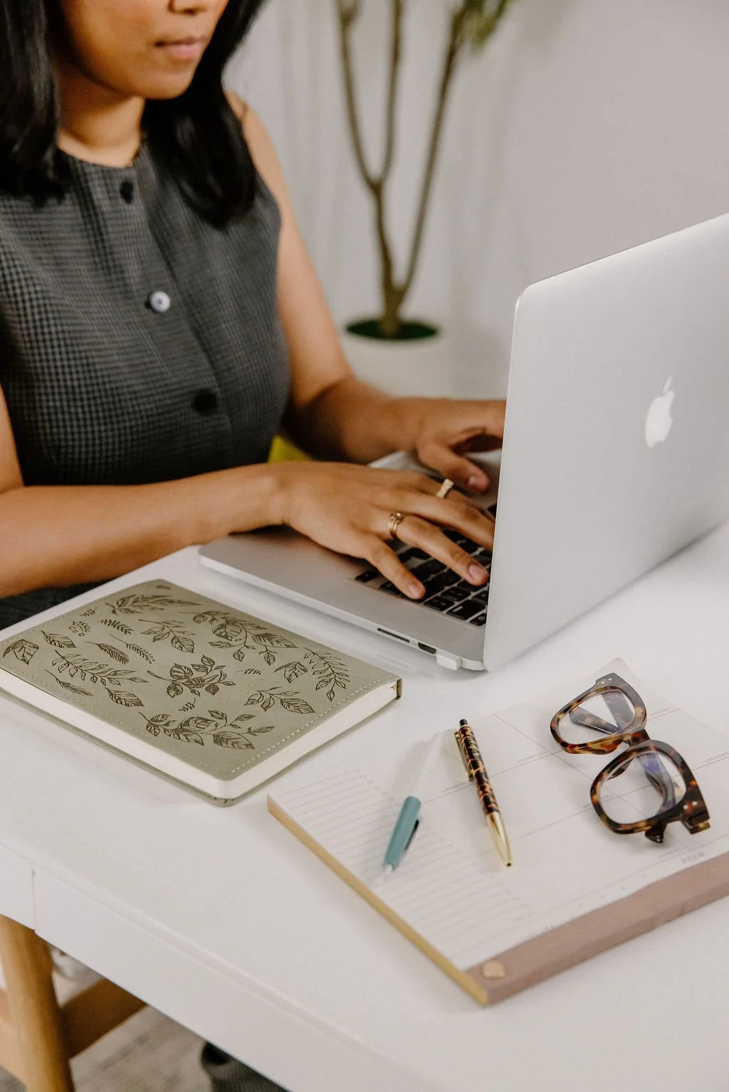 A woman typing on a silver Apple MacBook laptop on a white desk, with a notebook, pens, glasses, and note paper nearby, and a potted plant in the background.