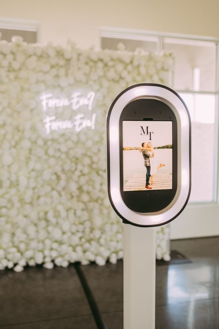 A love-themed photo booth with a screen showing a couple embracing and kissing on a dock, with white flowers and a neon sign in the background.