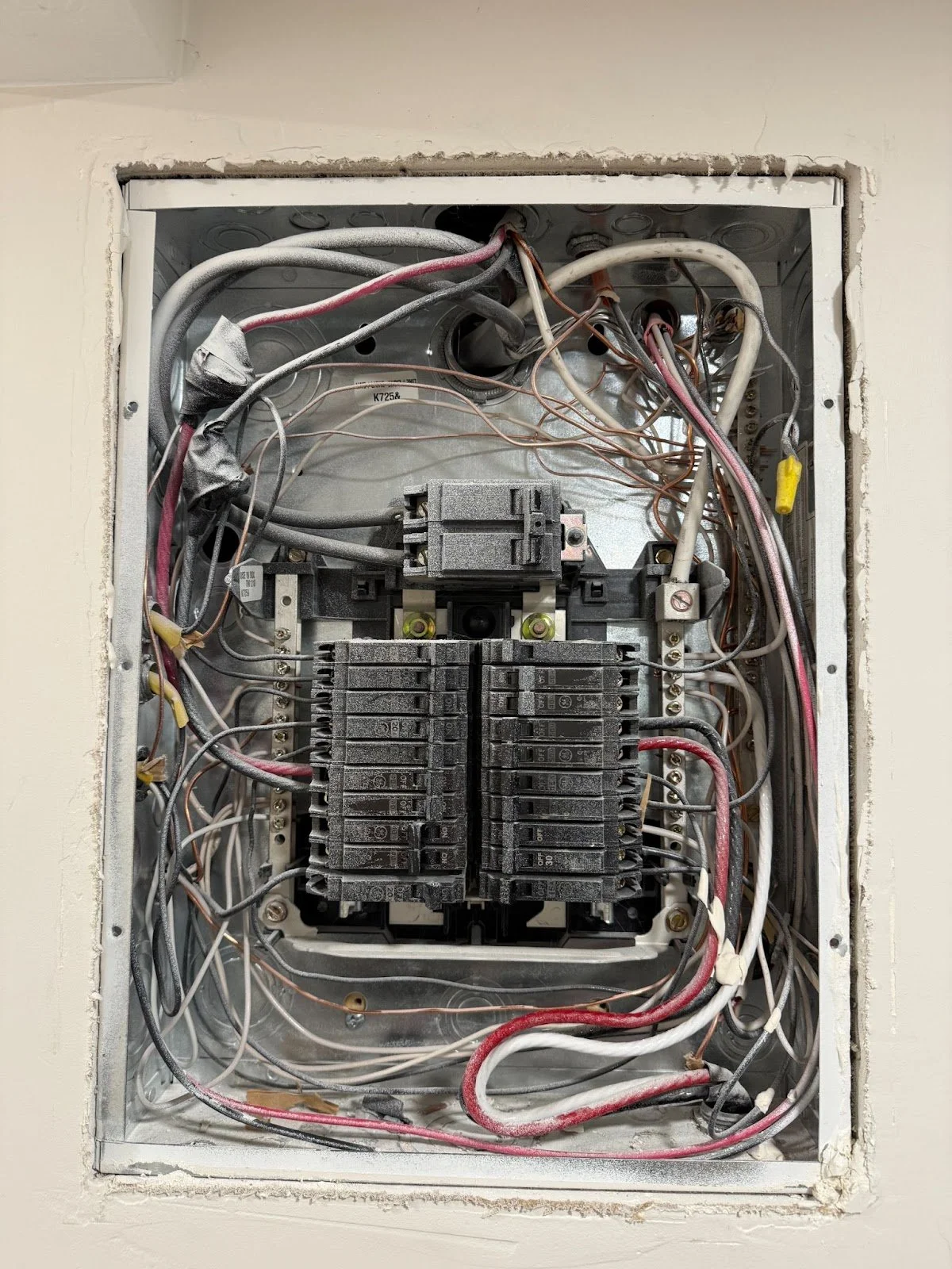 An open electrical panel box recessed in a wall, showing a complex arrangement of circuit breakers and dusty wiring.