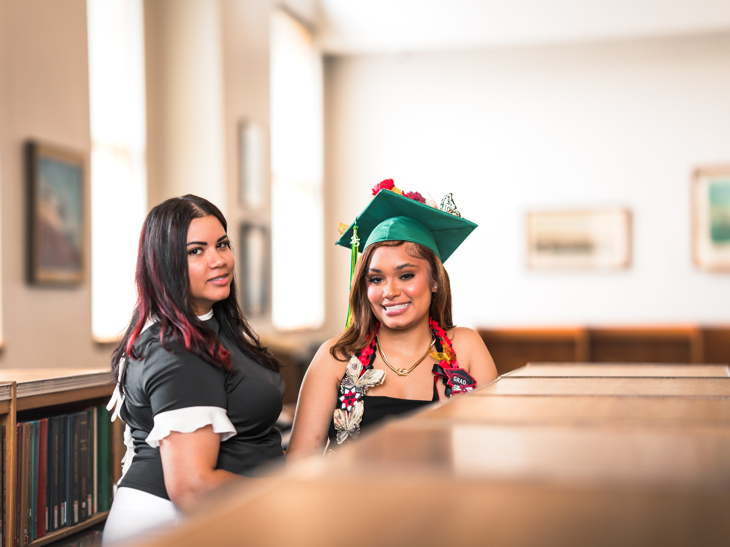 a young woman in graduation cap and gown smiling with another woman in a black uniform inside a library.