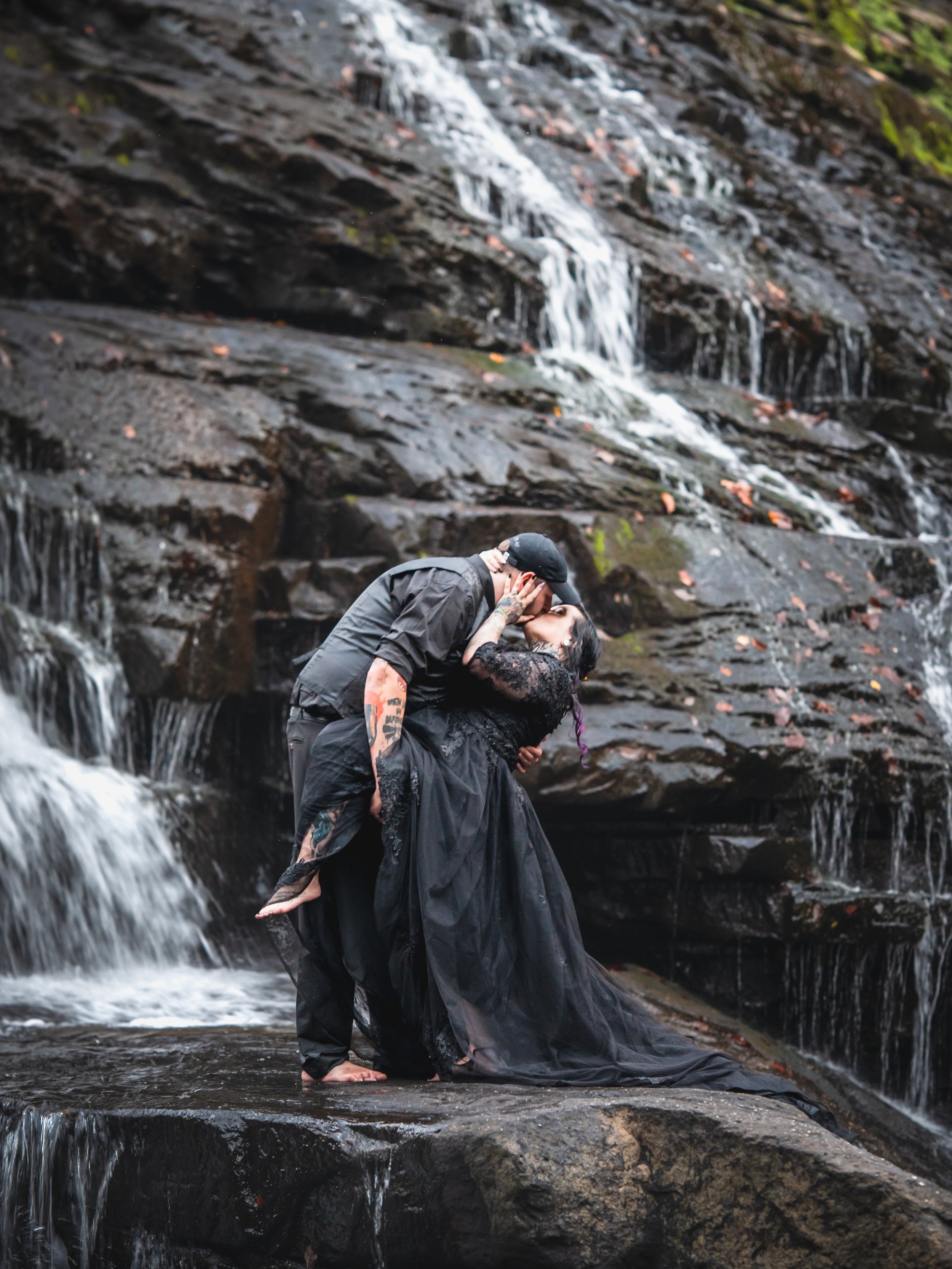 A couple is kissing on a wet rock in front of a small waterfall in a natural setting.
