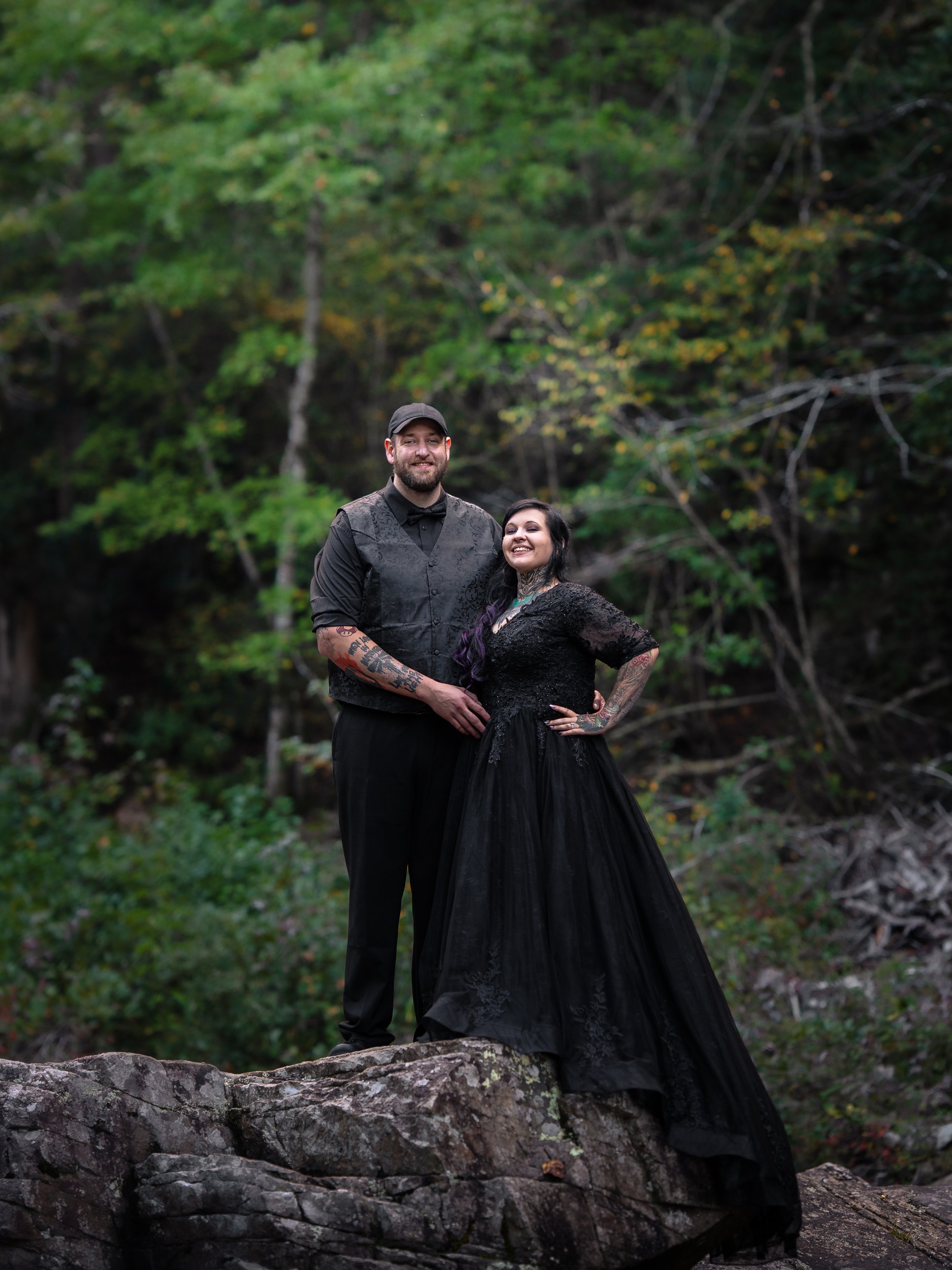 A couple dressed in black, standing on a large rock in a forested area, smiling and posing together.