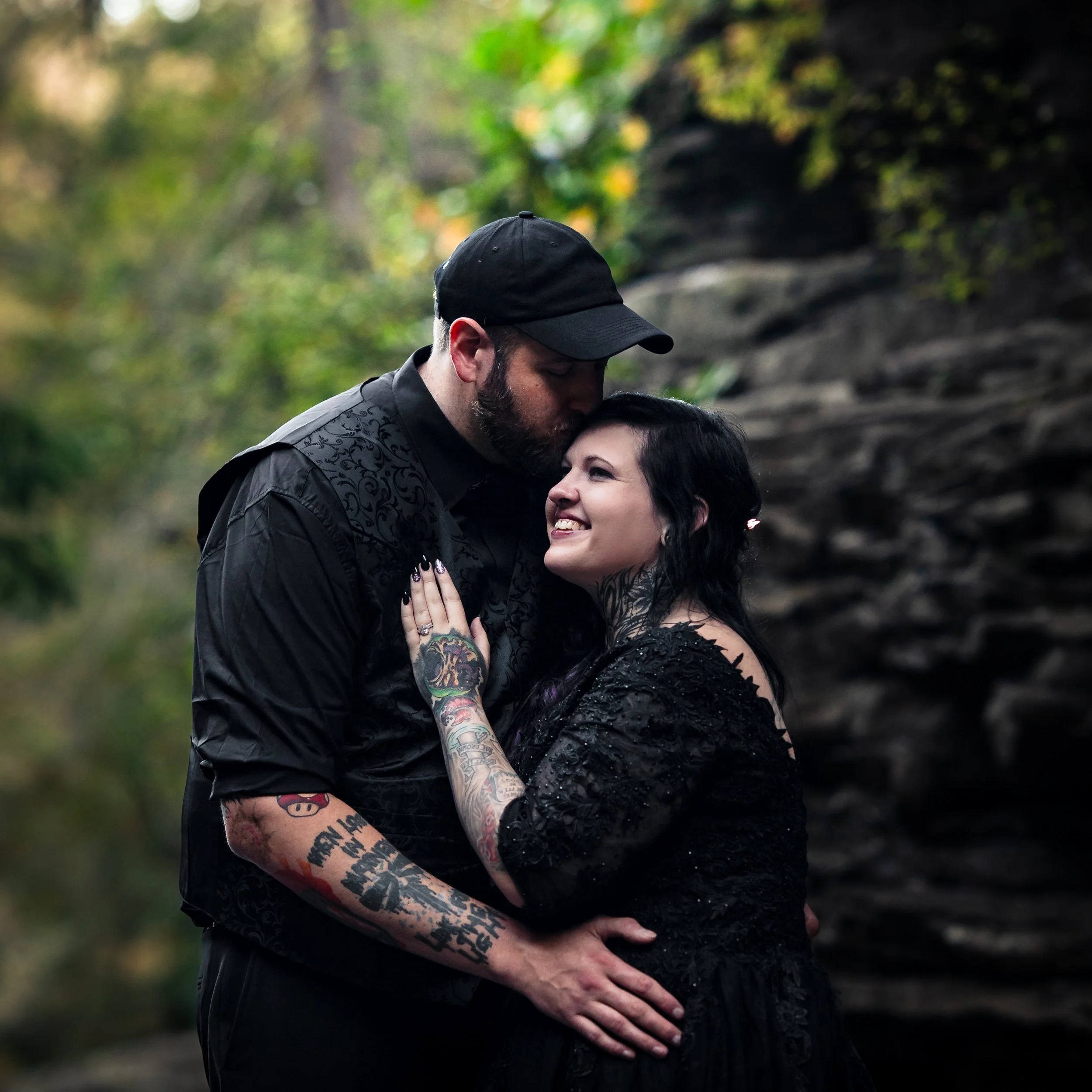 A couple embraces outdoors, with the man kissing the woman on the forehead. Both are smiling, and the woman has tattoos on her arms and neck. The background is blurred with greenery and rocks.