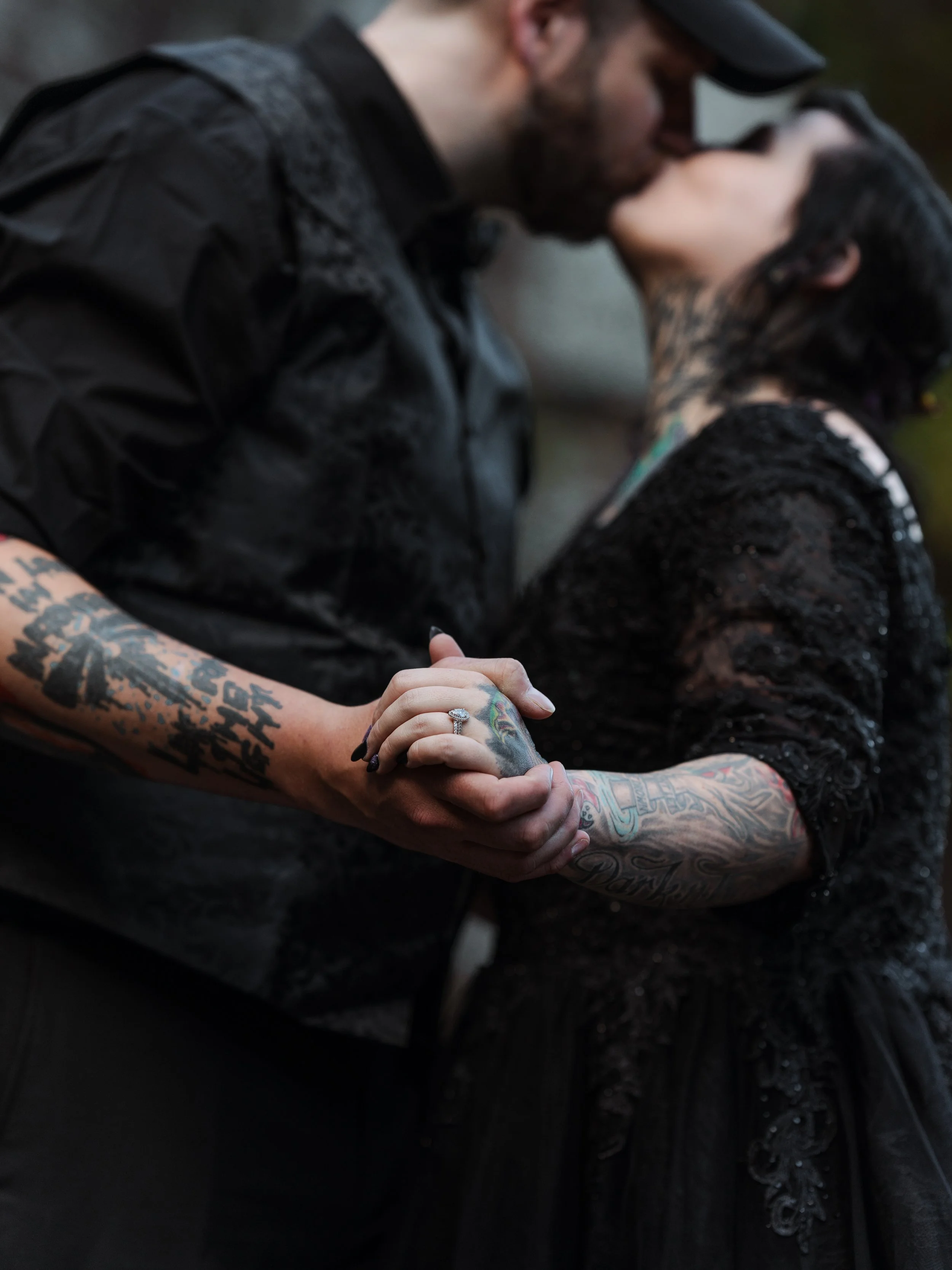 A couple is kissing and holding hands, with the man wearing a black jacket and cap and the woman wearing a black lace dress, both displaying tattoos on their arms.