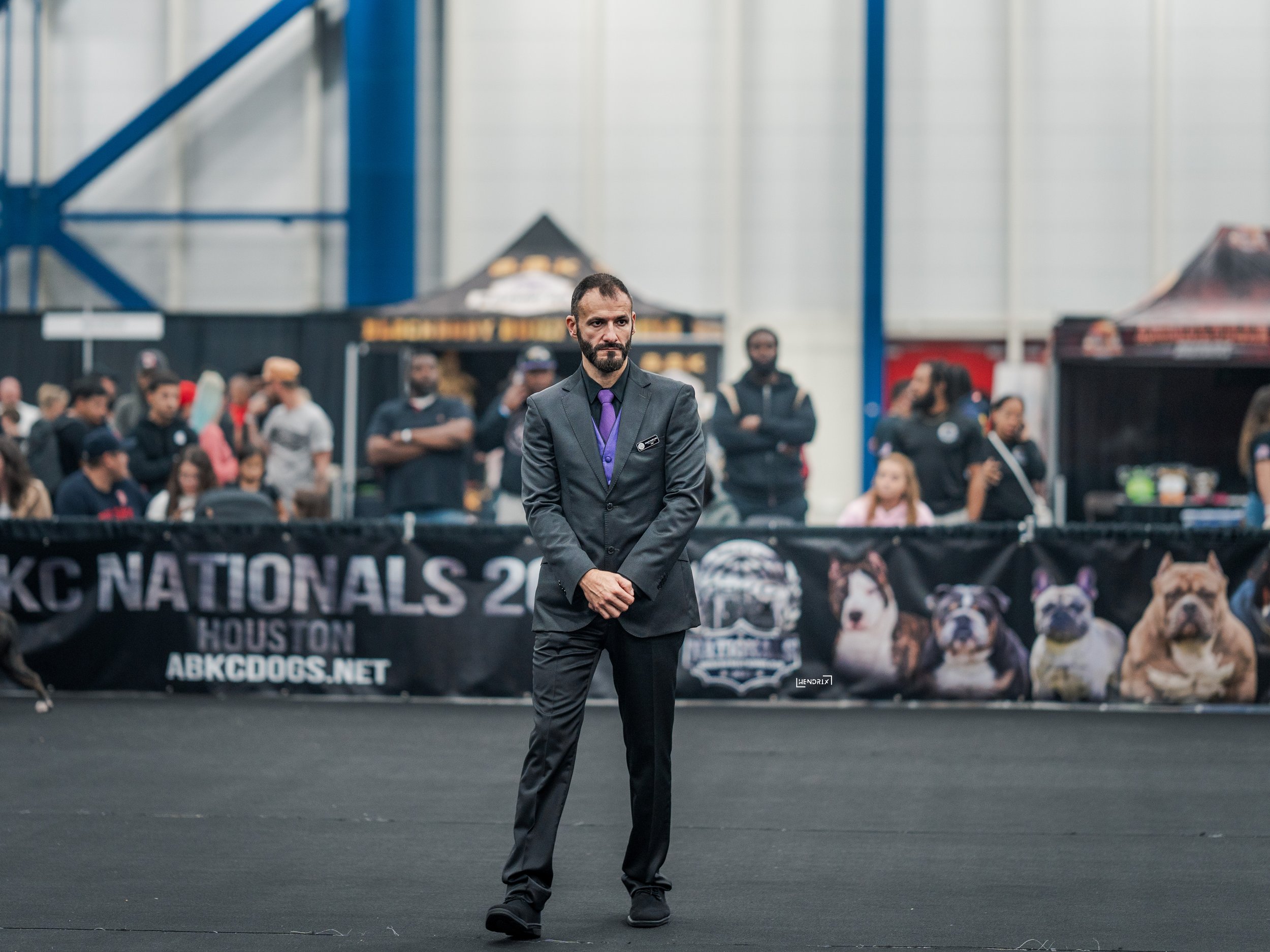 A man in a suit stands with crossed hands in front of a black banner at a dog show, with an audience and display booths in the background.