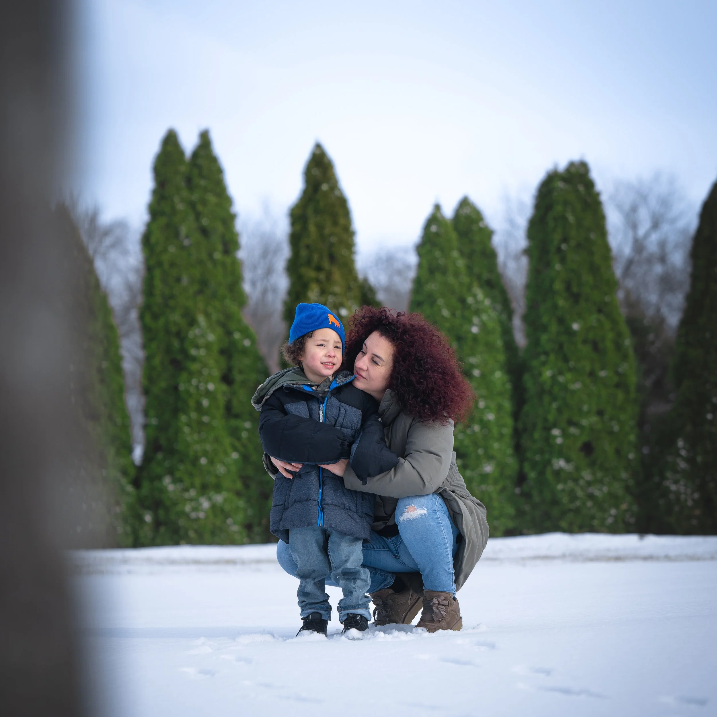Woman kneeling in snow, comforting crying boy wearing a blue beanie, in a winter outdoor setting with tall evergreen trees in background.
