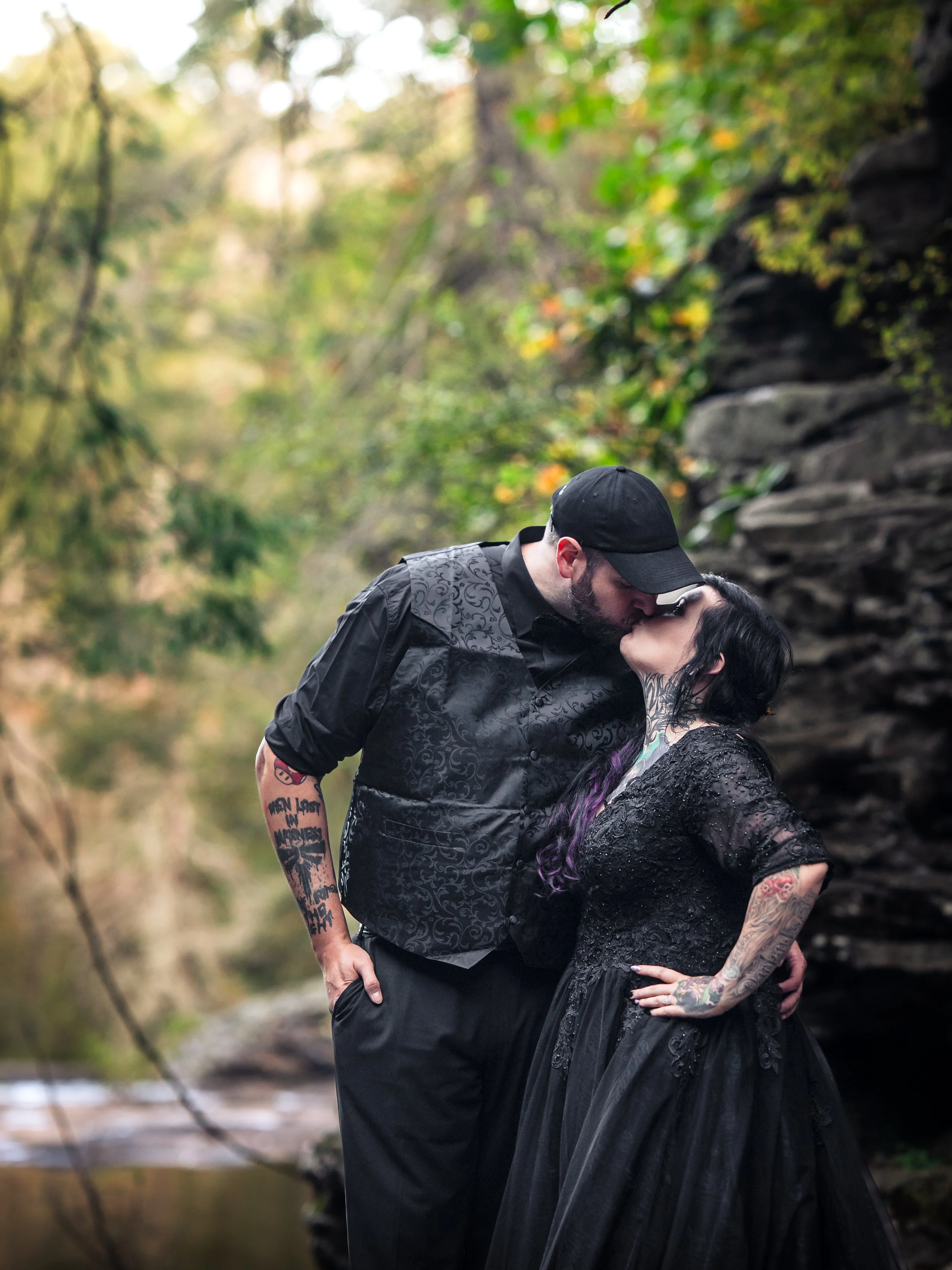 A couple kissing outdoors near a rocky cliff and a river, both dressed in dark clothing, with tattoos visible on their arms.