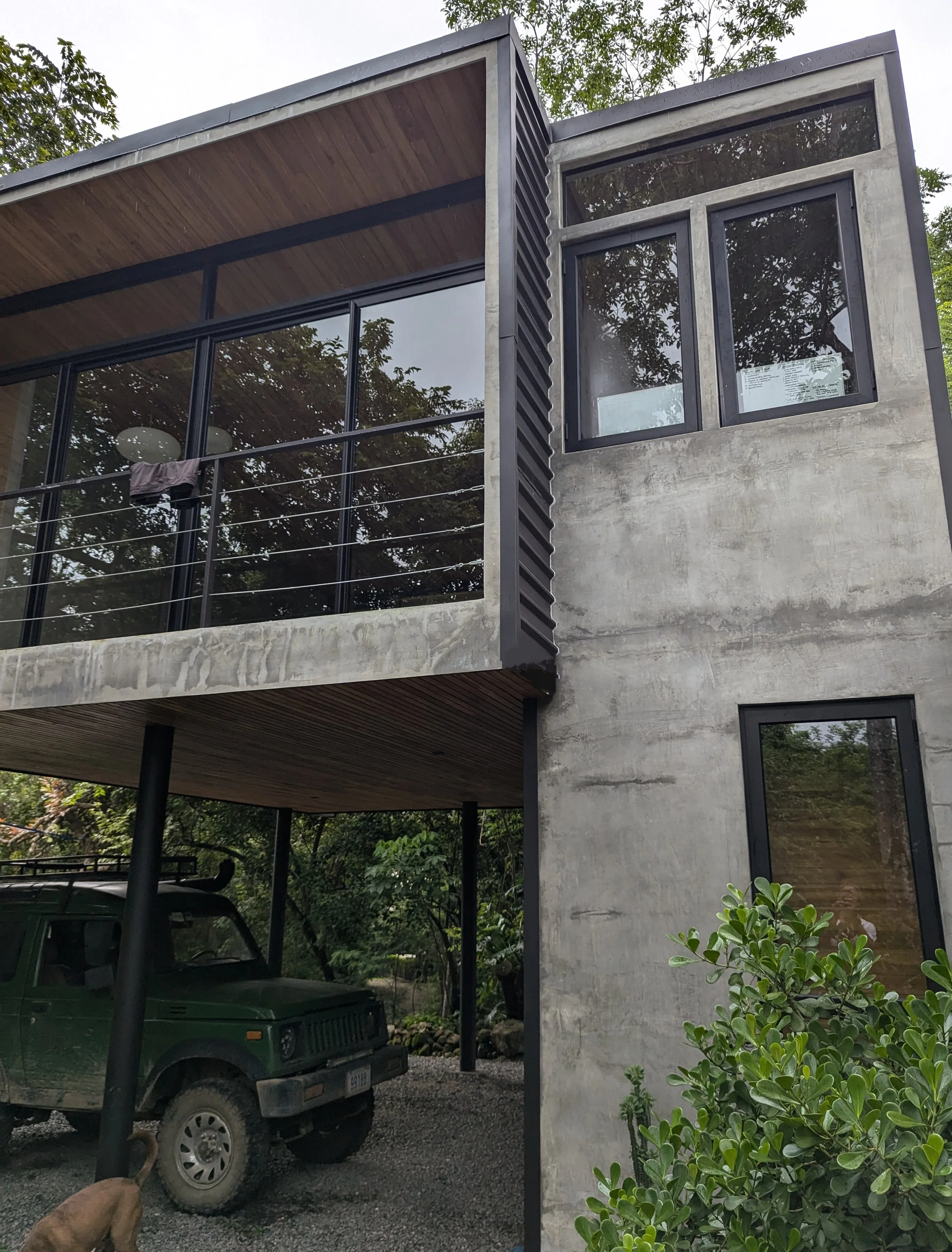 Modern multi-story house with concrete walls, large windows, and a balcony with glass railing. A green Land Rover is parked underneath the house, with a dog nearby.