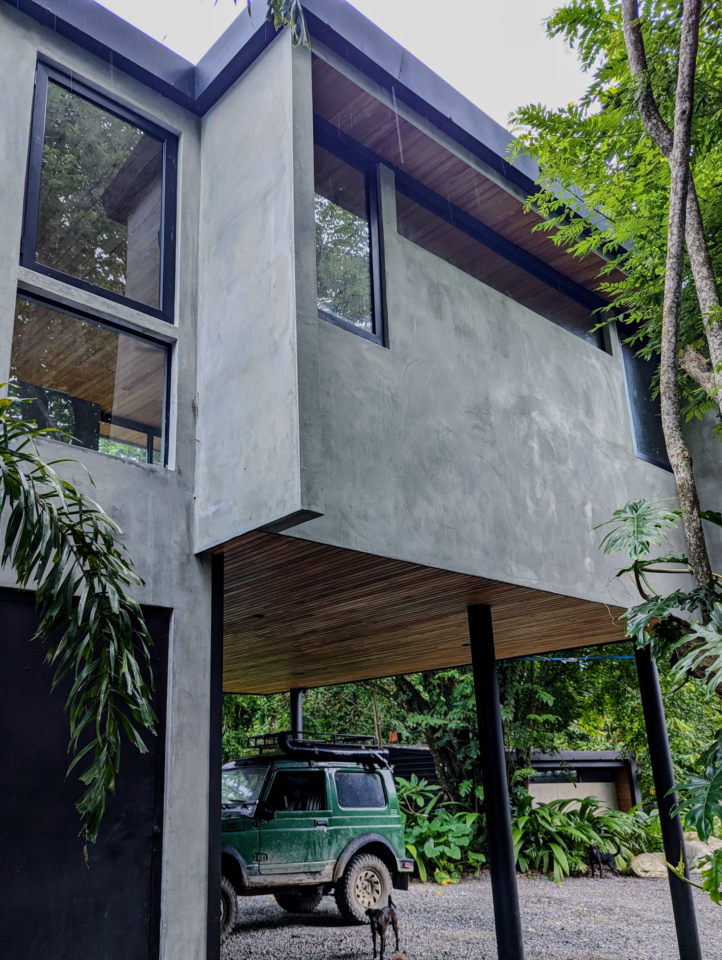 Modern house with concrete walls, large windows, wooden ceiling in carport area, surrounded by greenery, with a green off-road vehicle and a dog underneath the house.