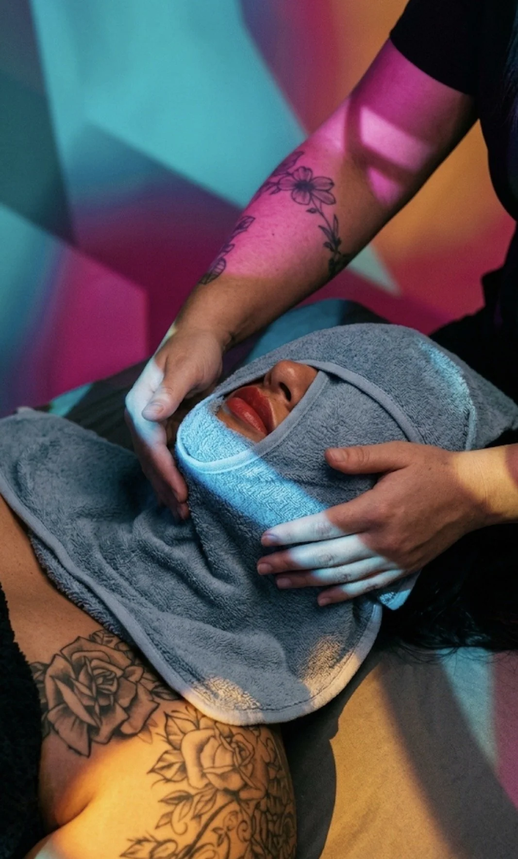 A person getting a facial massage, with their face wrapped in a towel and a towel covering their head, while a massage therapist with tattoos receiving colorful lighting performs the treatment.