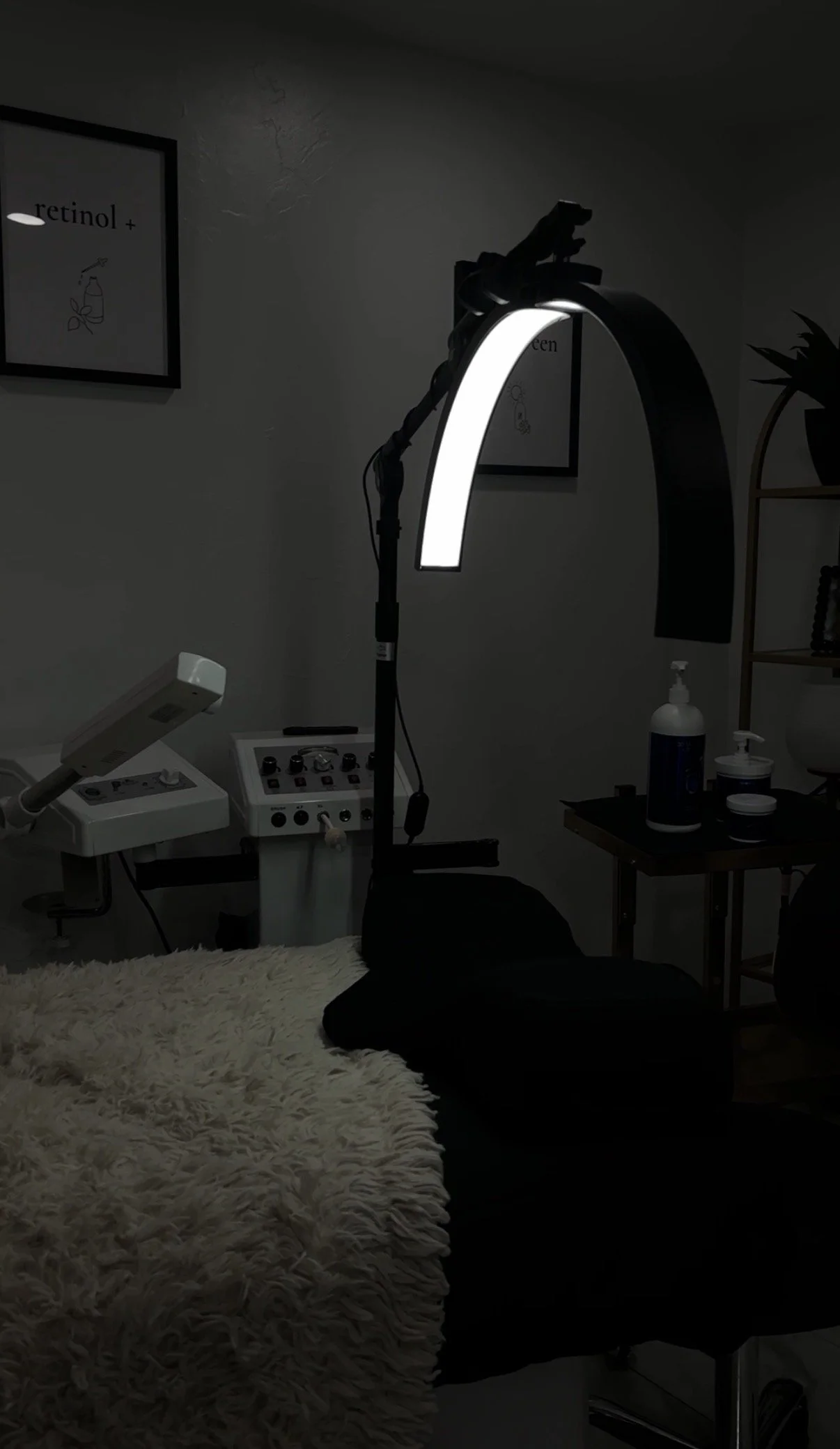 Dark room with medical or aesthetic equipment, including a large curved light, a framed poster on the wall with the text 'retinol+', a fluffy rug, and bottles of skincare or medical supplies on a small table.