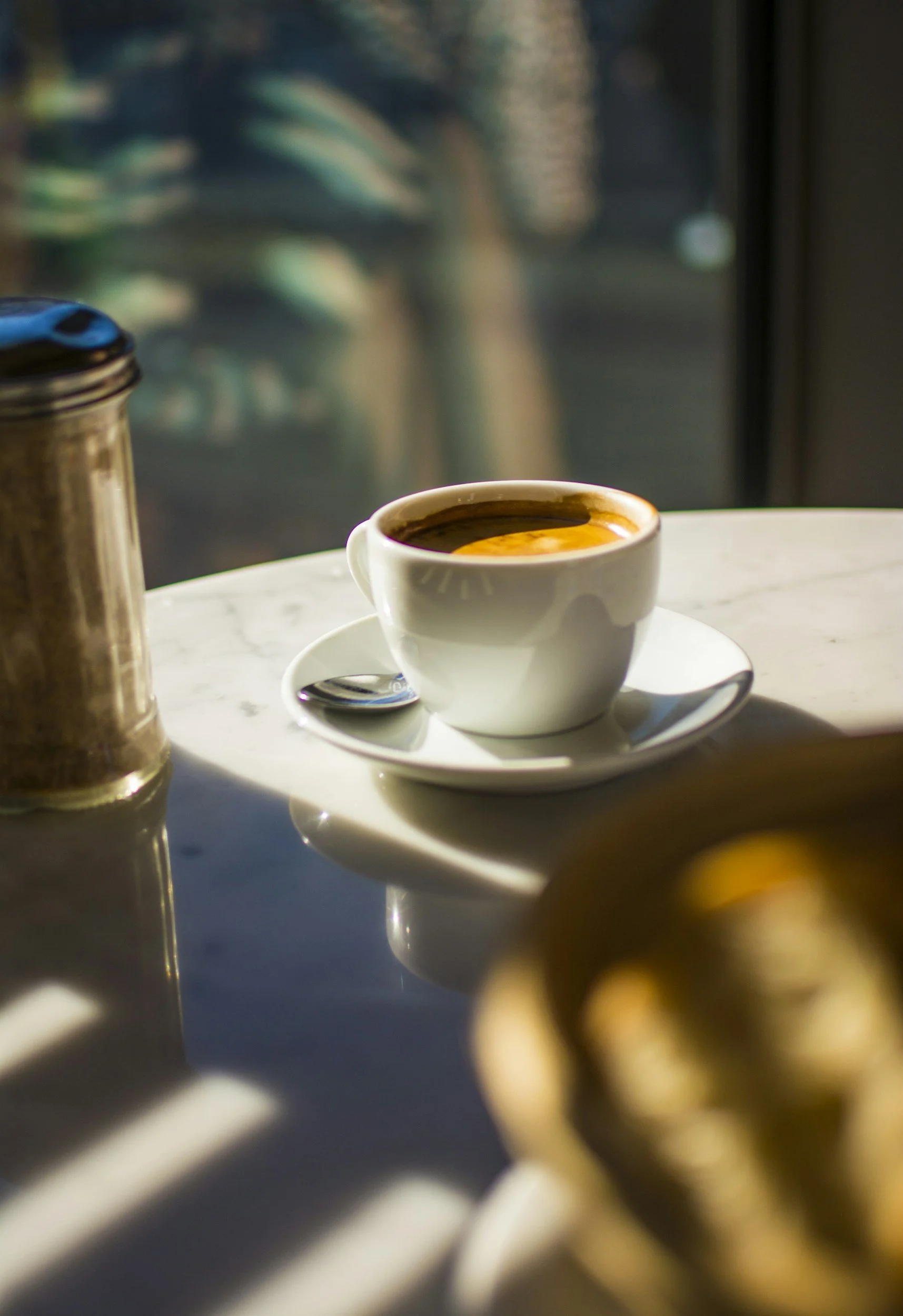 A white cup filled with coffee on a saucer with a small spoon, sitting on a white marble table with sunlight and shadows.