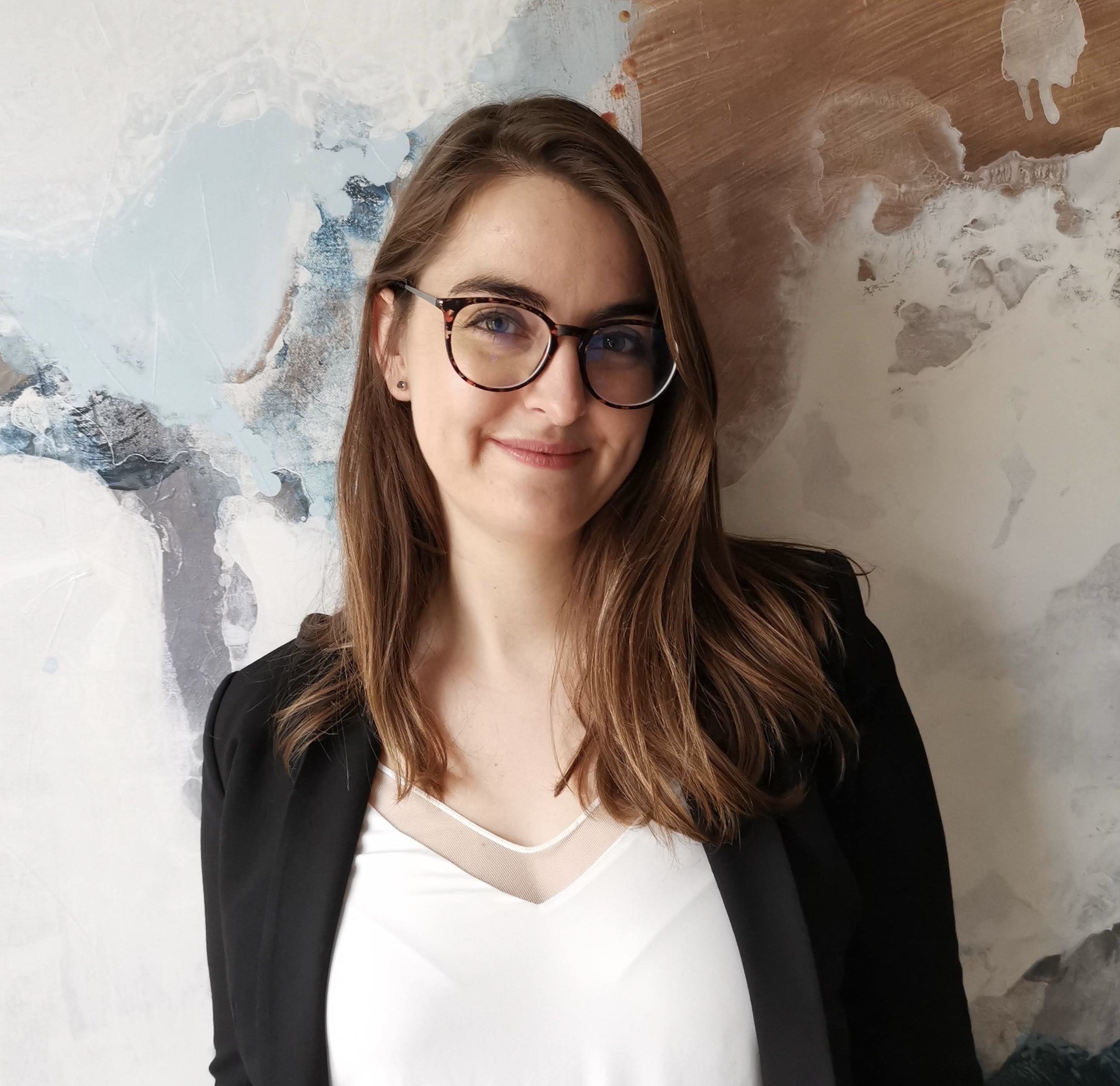 A woman with long brown hair, glasses, and a slight smile standing against a textured abstract wall background.