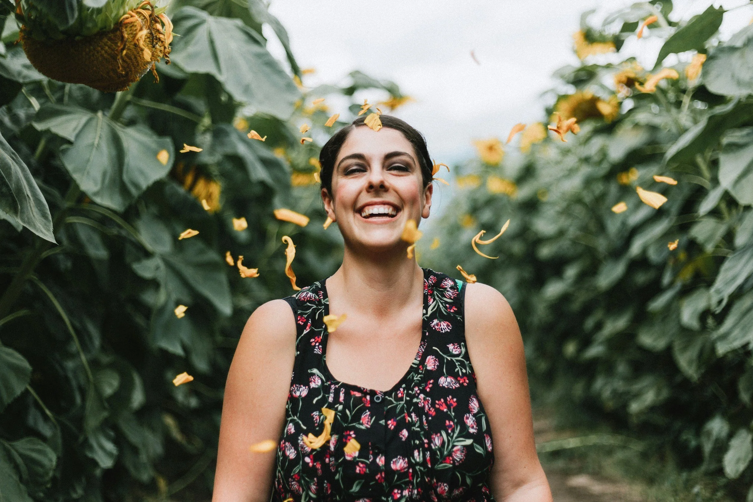 A woman smiling and throwing yellow flower petals in a sunflower field during daytime.