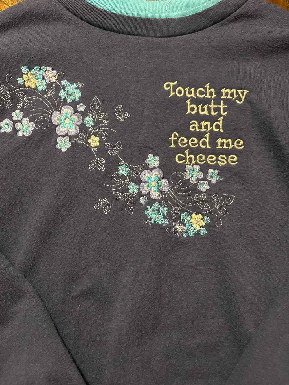 A black sweatshirt with embroidered floral patterns and yellow text that reads, "Touch my butt and feed me cheese."