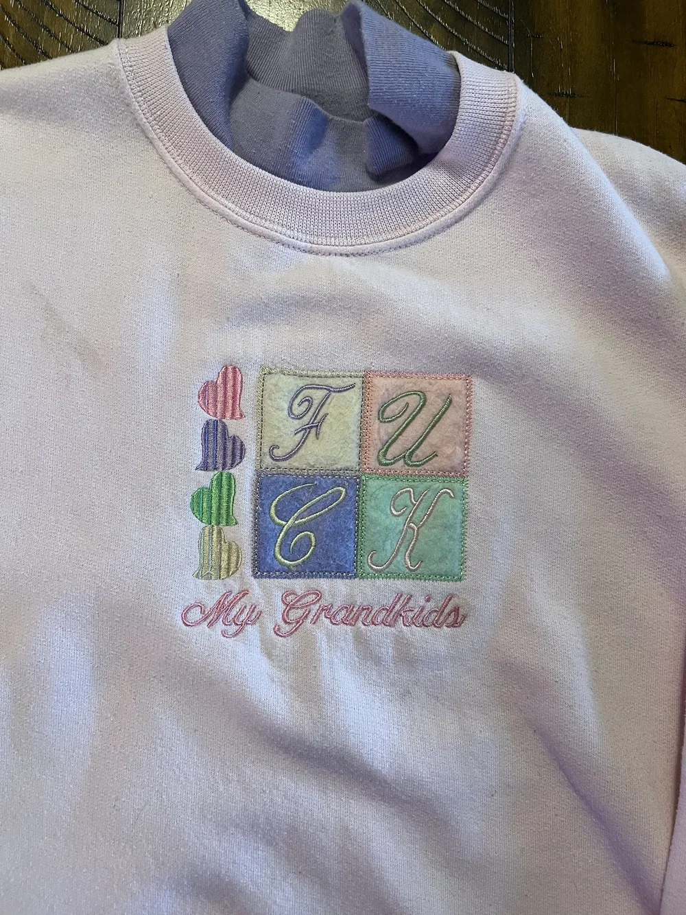 White sweatshirt with colorful embroidered design of four squares containing cursive letters 'F', 'U', 'C', 'K' and a row of four small embroidered hearts next to the squares. Text below says 'My Grandkids' in pink cursive.