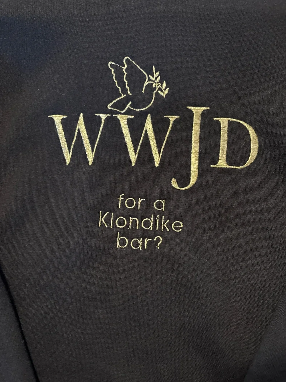 Black fabric with embroidered text that reads "WWJD" and "for a klondike bar?" with a simple outline of a dove holding an olive branch above the text.