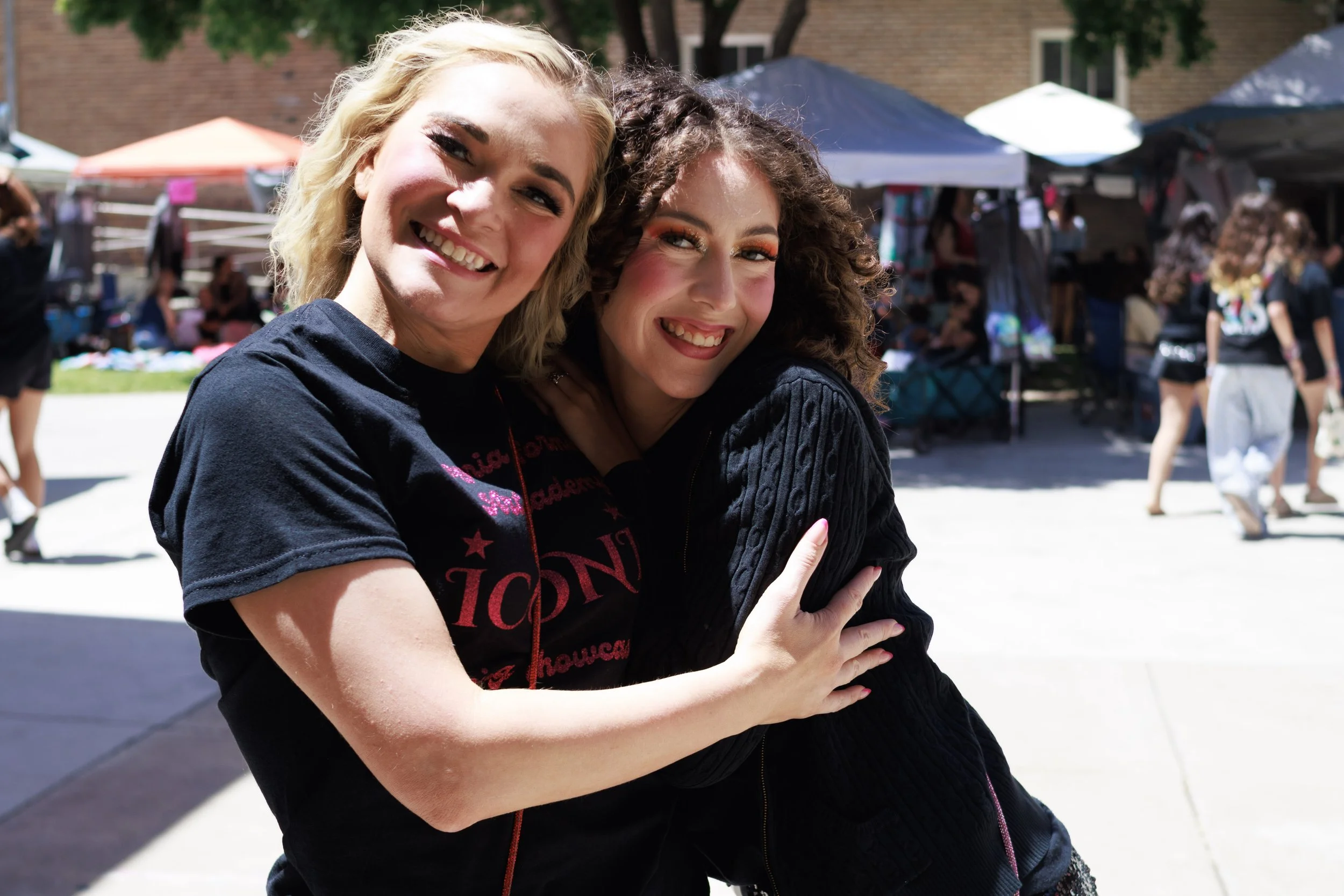 Two women hugging and smiling at an outdoor event with tents and people in the background.