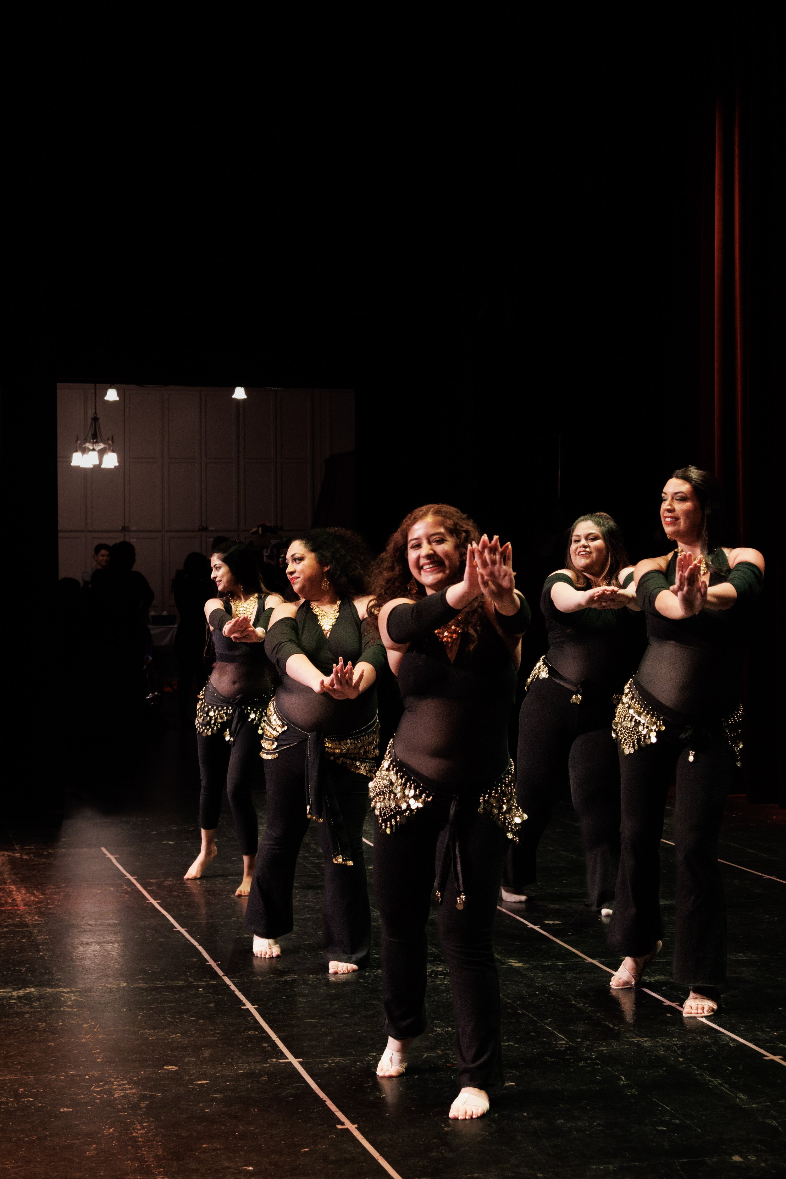 Group of adults performing a dance on stage, dressed in black costumes with gold embellishments, smiling and clapping in unison.