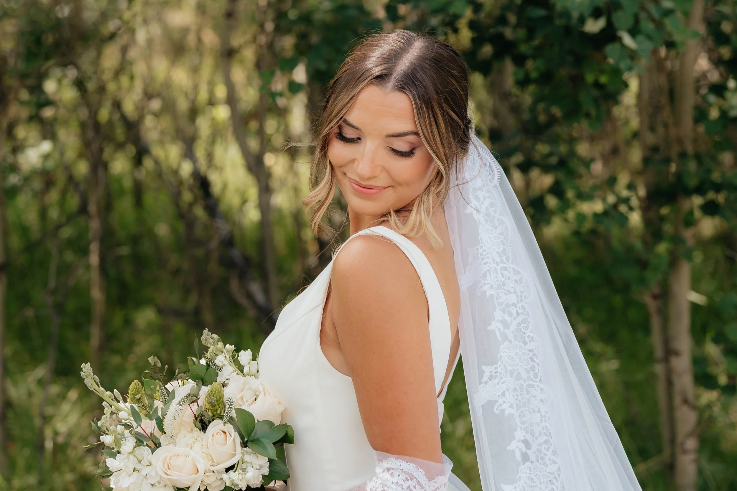 Close-up of a smiling woman with blonde hair and eyes closed, wearing a white dress with a beaded strap, outdoors with a blurred background.