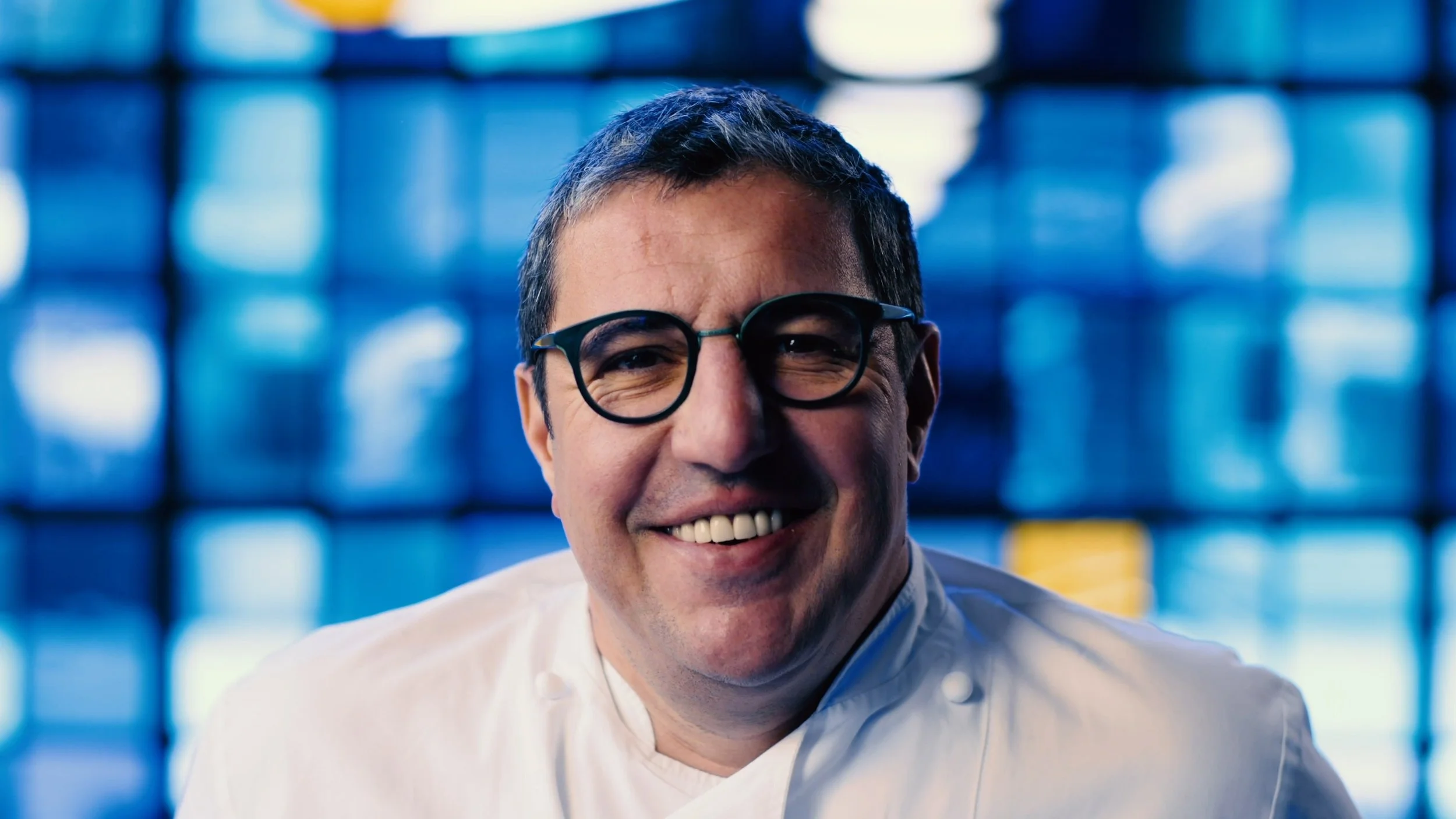 A smiling man with glasses wearing a white shirt, standing in front of blue illuminated panels.