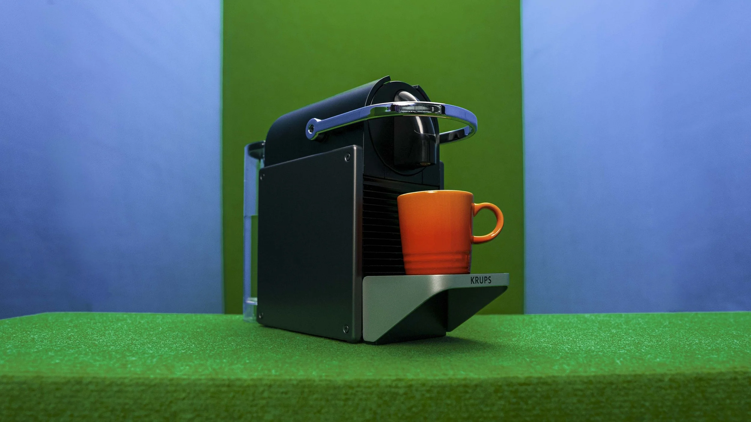 A small black coffee machine with a brand label 'KRUPS' on it, placed on a green surface against a background with blue and green sections. An orange coffee mug is positioned underneath the dispenser of the coffee machine.