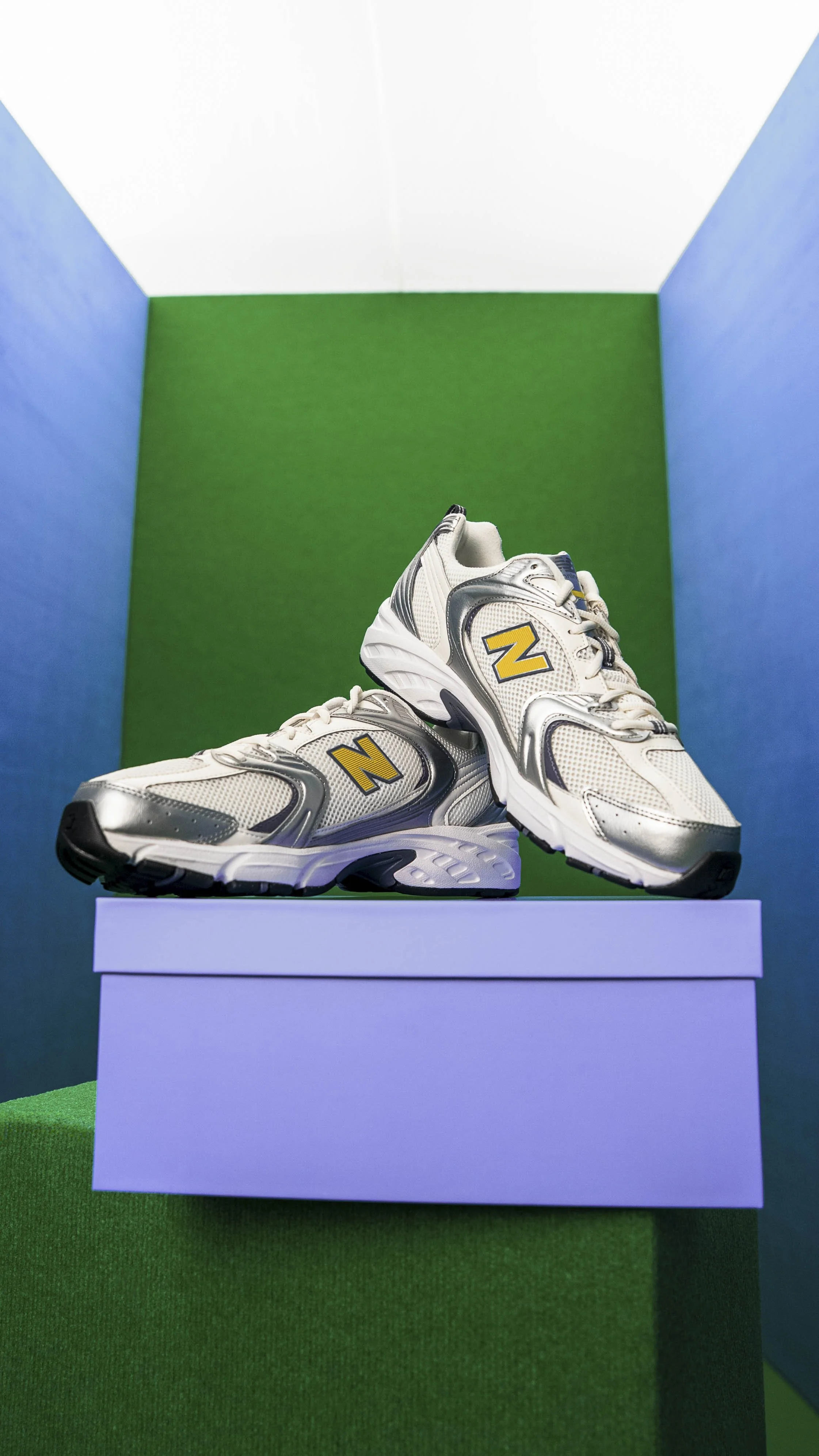 Pair of white athletic shoes with silver and yellow accents, displayed on a white platform against a green and blue background.
