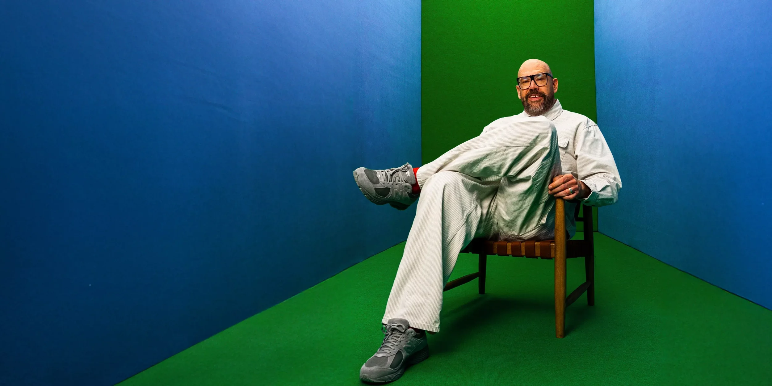 A man with glasses and a beard sitting on a wooden chair in a room with blue and green walls. He is wearing a light-colored outfit, sneakers, and red socks, holding one foot up on his knee.