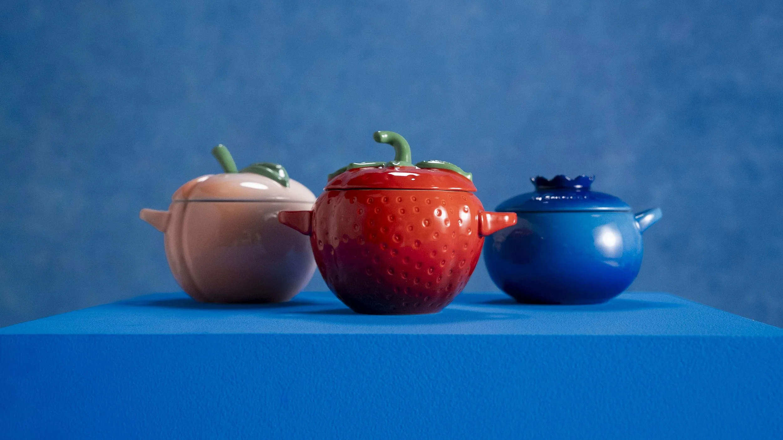 Three colorful strawberry-shaped teapots with lid handles resembling strawberries and leaves, placed on a blue surface against a matching blue background.