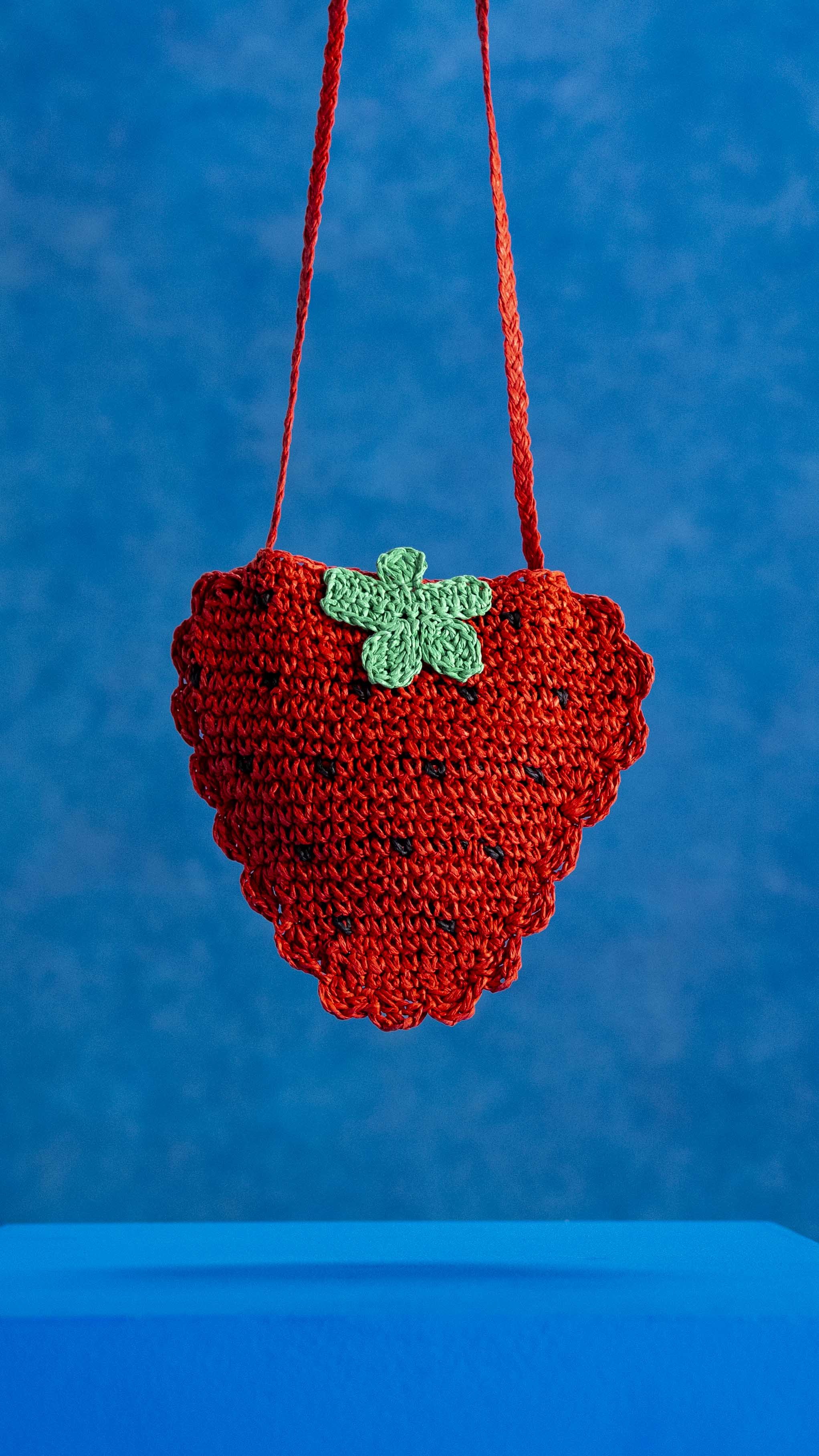 Crocheted strawberry-shaped purse with a green leaf detail, hanging by two red straps against a blue background.