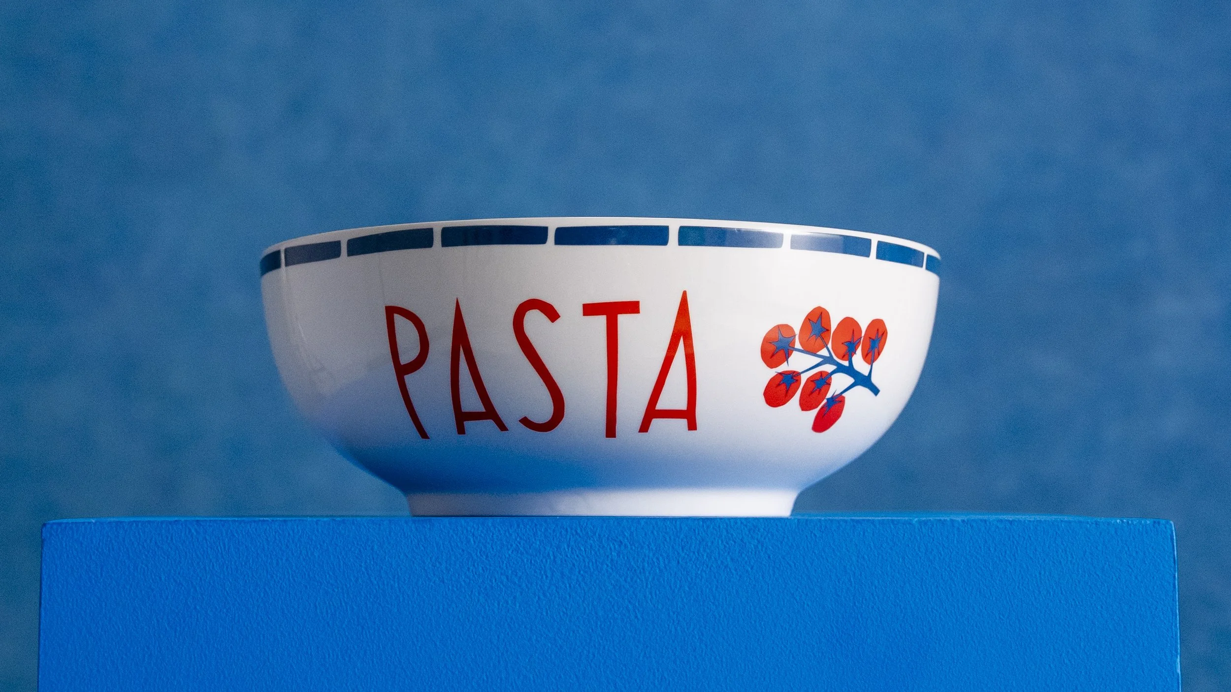 A white bowl with the word 'PASTA' in red and a blue and orange floral design, sitting on a blue surface against a blue background.