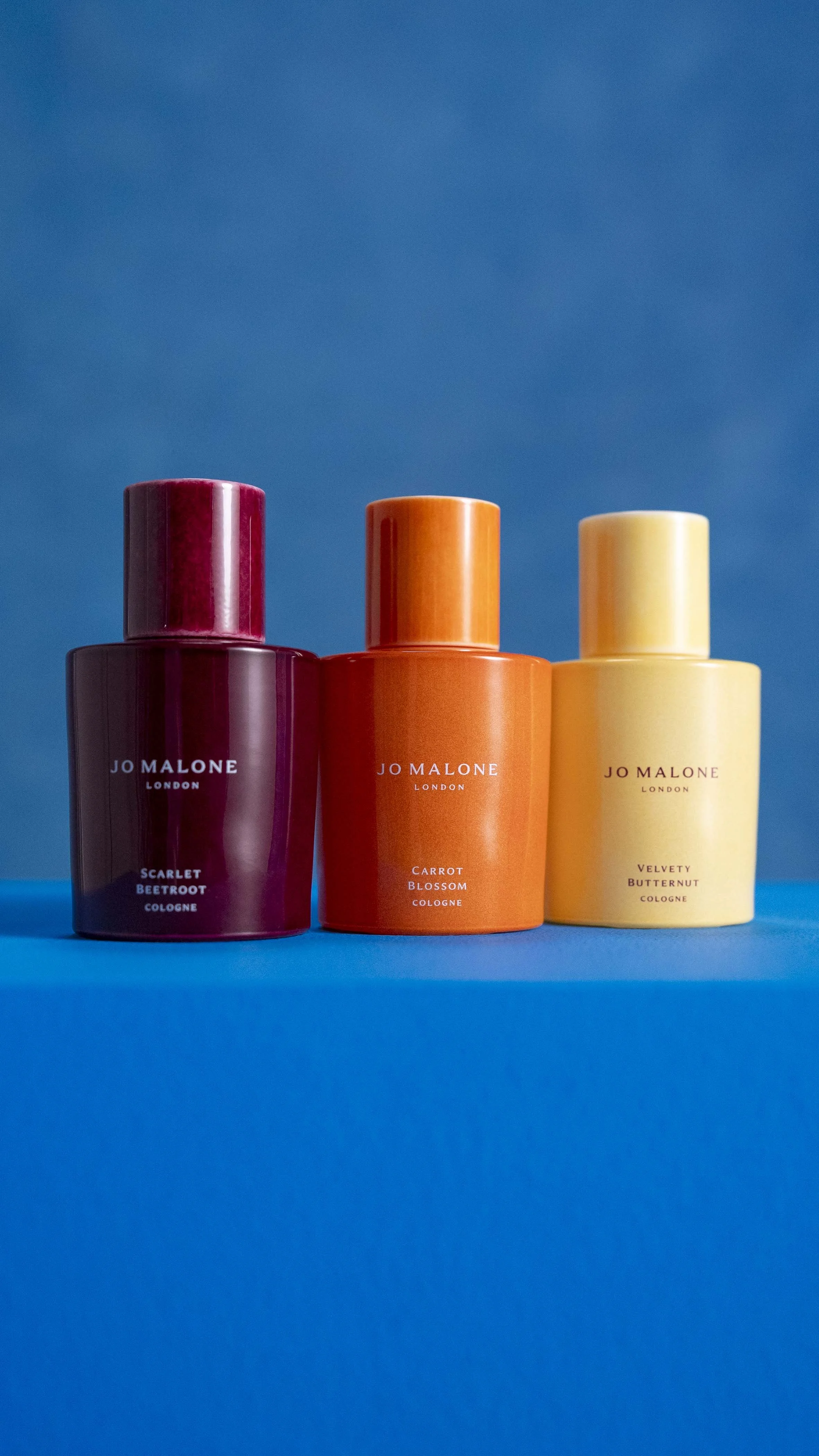 Three Jo Malone cologne bottles labeled Scarlet Beetroot, Carrot Blossom, and Velvety Butternut standing against a blue background.