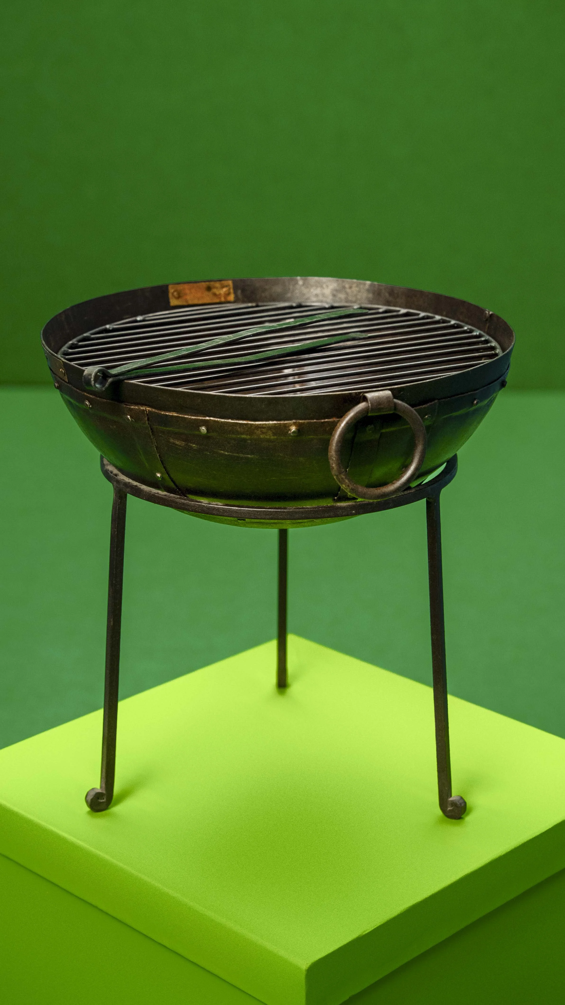 A vintage round metal grill with a handle and legs, displayed on a bright green platform against a green background.