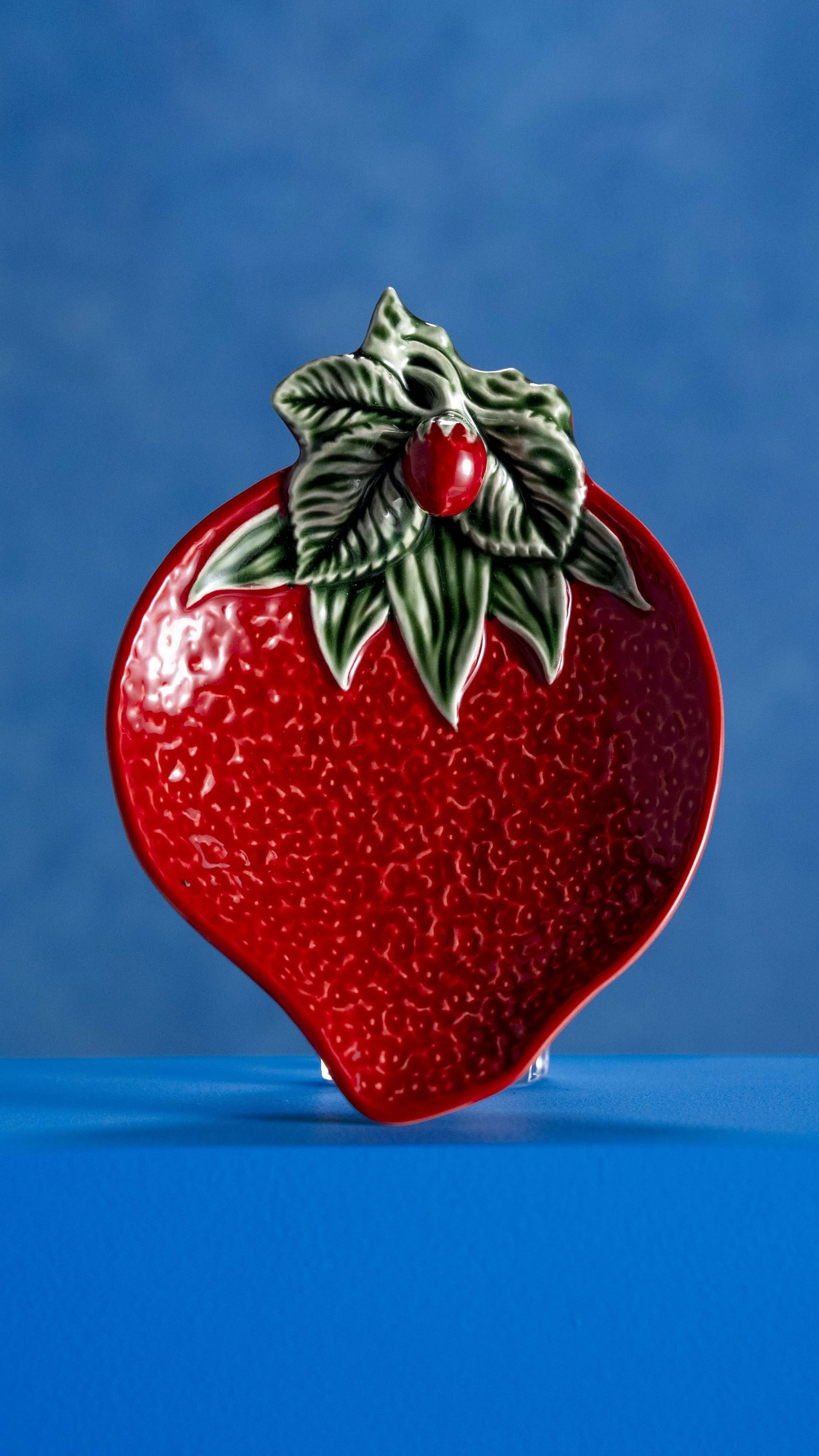 A decorative ceramic dish designed to look like a strawberry, with green leaves and a red berry on top, against a blue background.