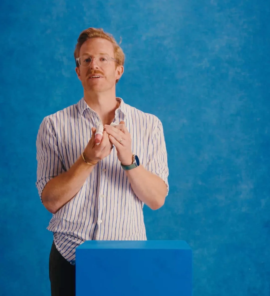 A man with glasses, red hair, and a beard, wearing a striped shirt, stands in front of a blue background. He is holding his collar with both hands and appears to be speaking.
