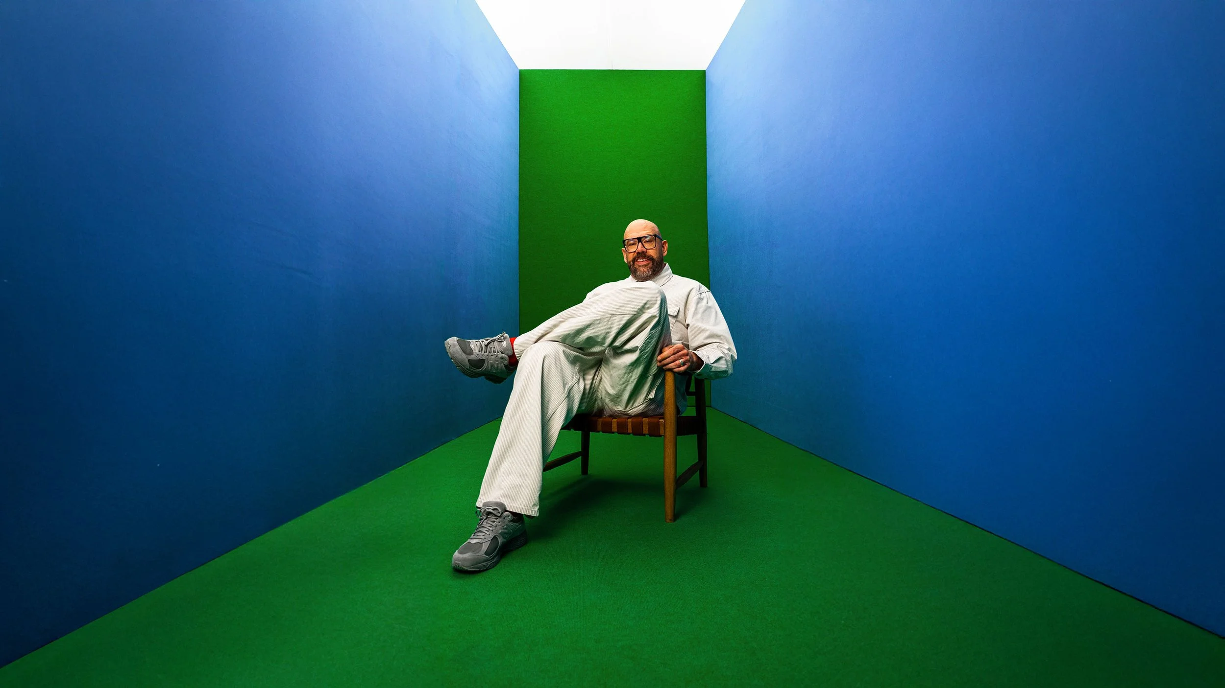 A man with glasses, a beard, and bald head sitting on a wooden chair in a small room with blue walls and a green floor and back wall. He is wearing a light gray tracksuit and gray sneakers, with one leg crossed over the other and smiling at the camera.