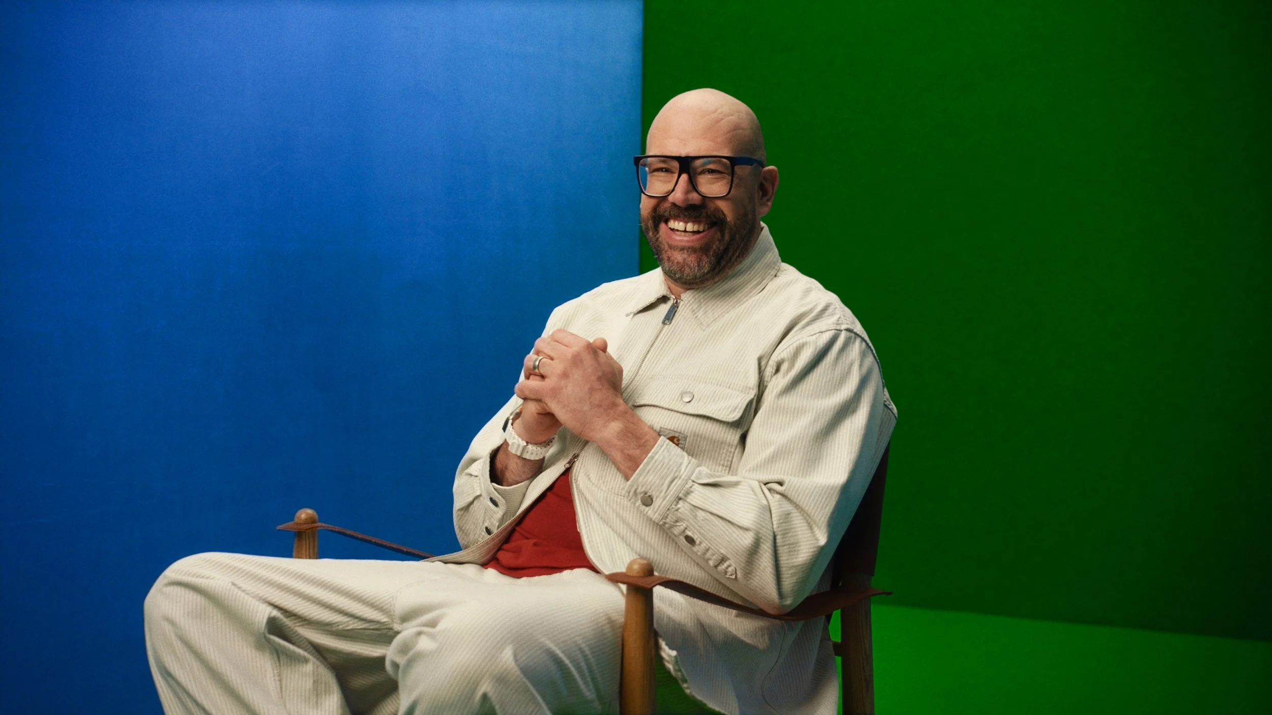 A smiling man with glasses and a beard sitting in a chair against a background divided into blue and green sections. He is wearing a beige jacket and trousers with a red shirt underneath.