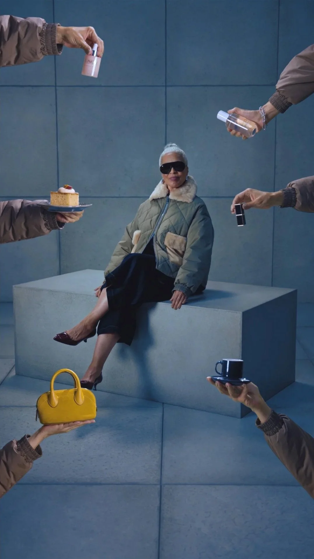 A woman with gray hair, sunglasses, and a fur-collared jacket sitting on a concrete block, surrounded by hands holding various objects like a cake, handbag, nail polish, bottle, mug, and lipstick, against a blue tiled wall.