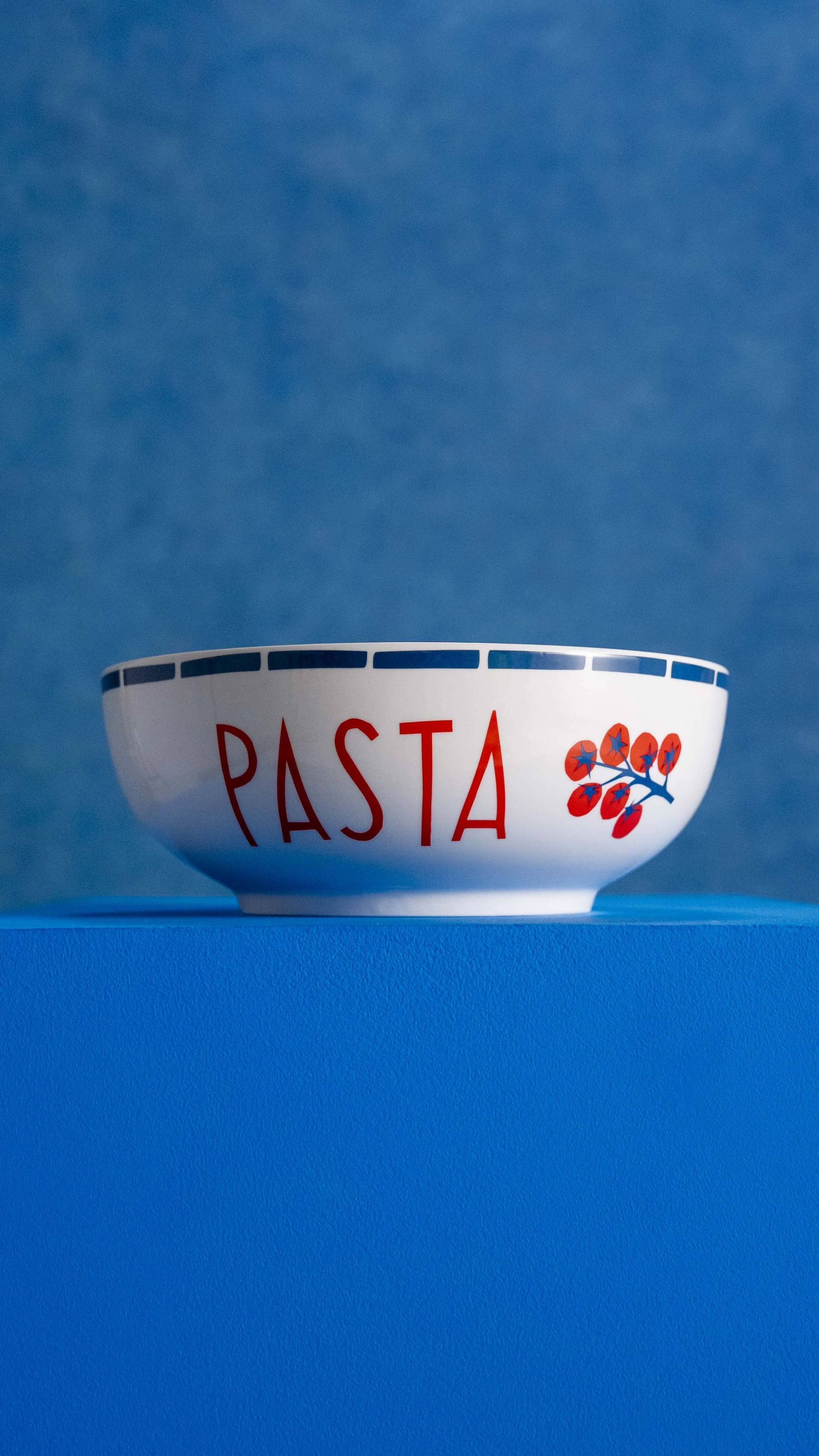A white bowl with the word 'PASTA' in red letters and a red berry branch illustration, set against a blue background.