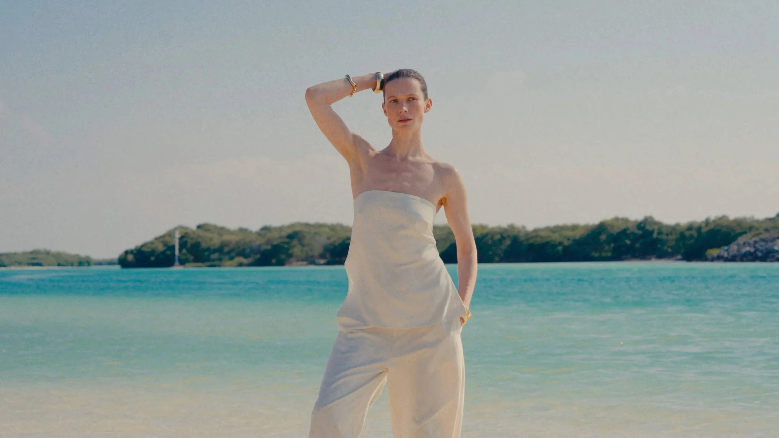 A woman standing on a beach with turquoise water and an island in the background, wearing a strapless white dress and jewelry, with her left hand on her head and looking into the distance.