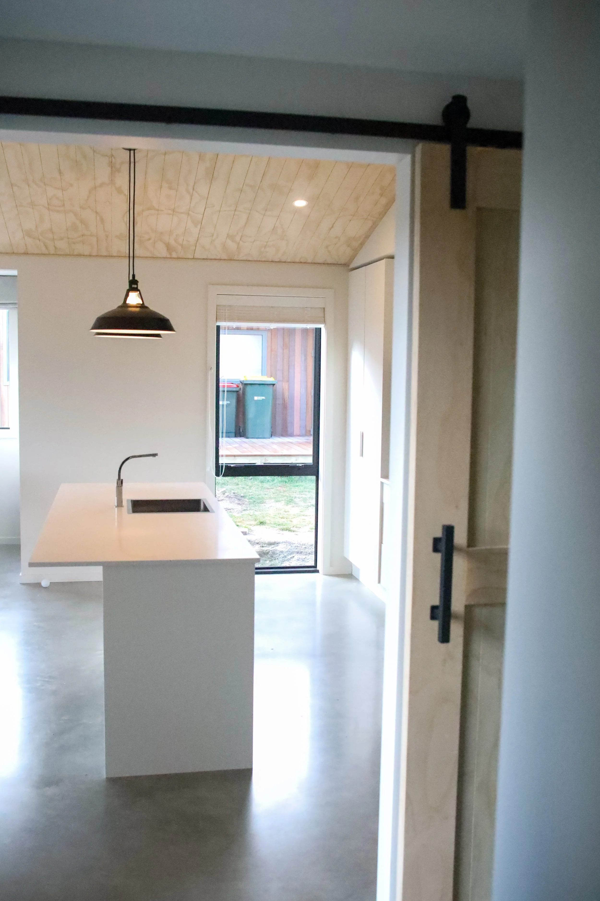 Modern kitchen with a white island, black sink, pendant light, and sliding glass door leading outside to a small yard with two trash bins.