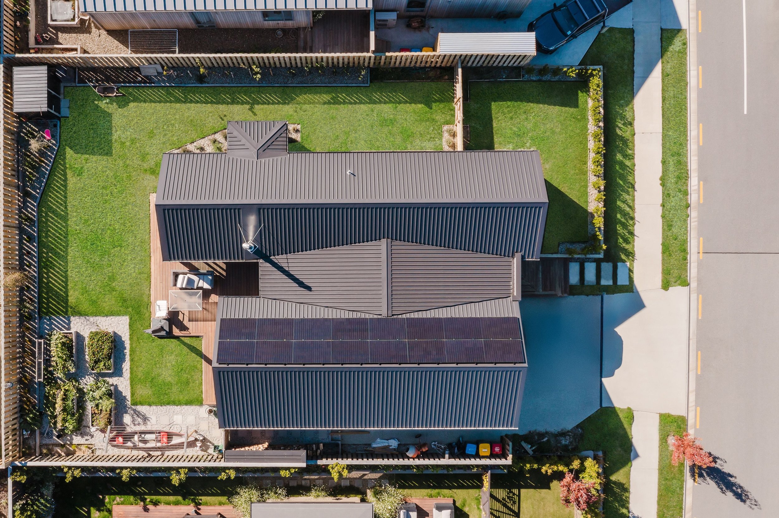Aerial view of a modern house with a dark metal roof, a small backyard with a lawn, a patio, and a solar panel on the roof. The house is adjacent to a street with a driveway and sidewalk, and is surrounded by a wooden fence and neighboring properties