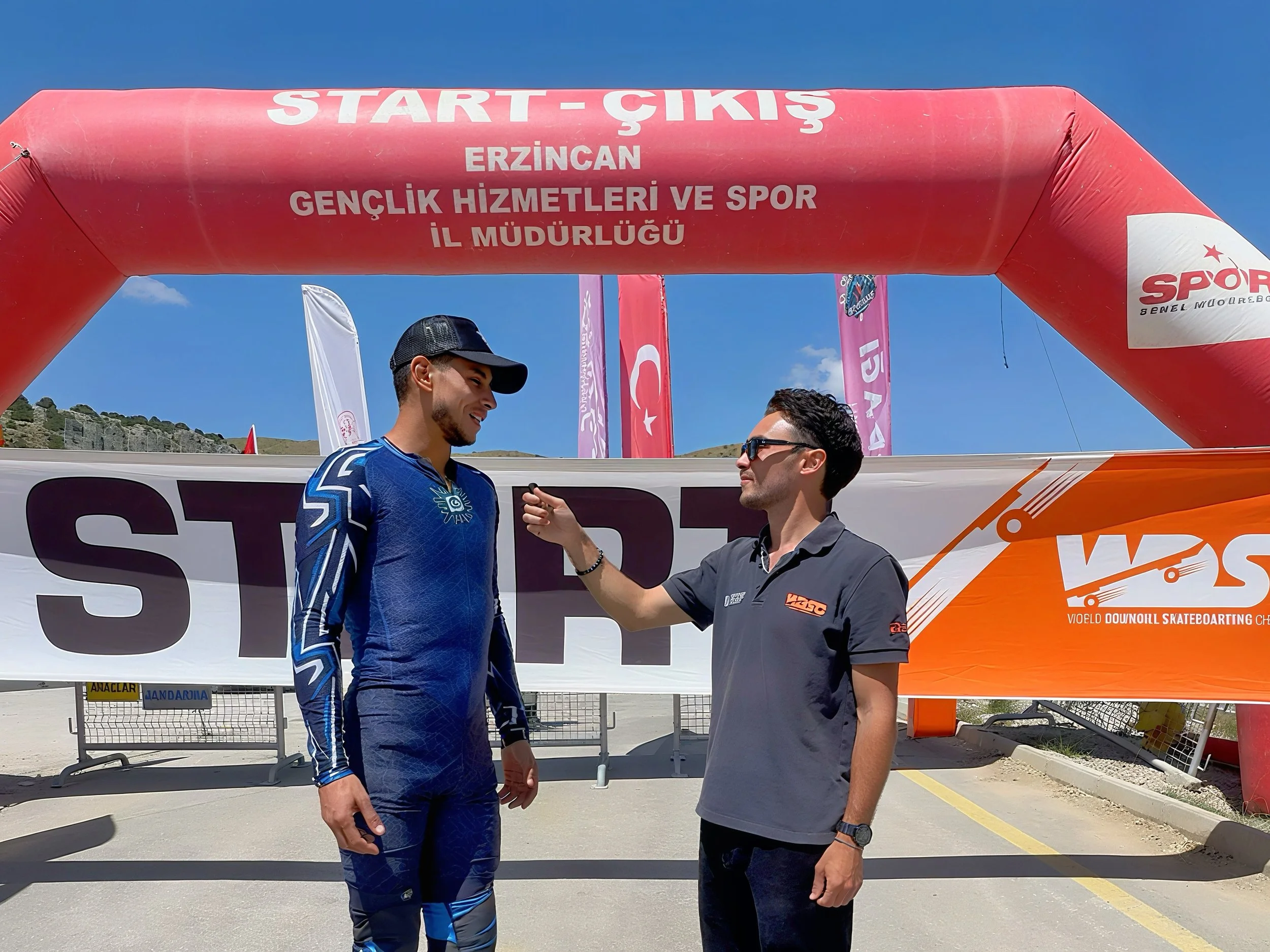 Two men speaking under a pink inflatable arch at a starting line for a sports event, with banners and a mountainous background on a sunny day.