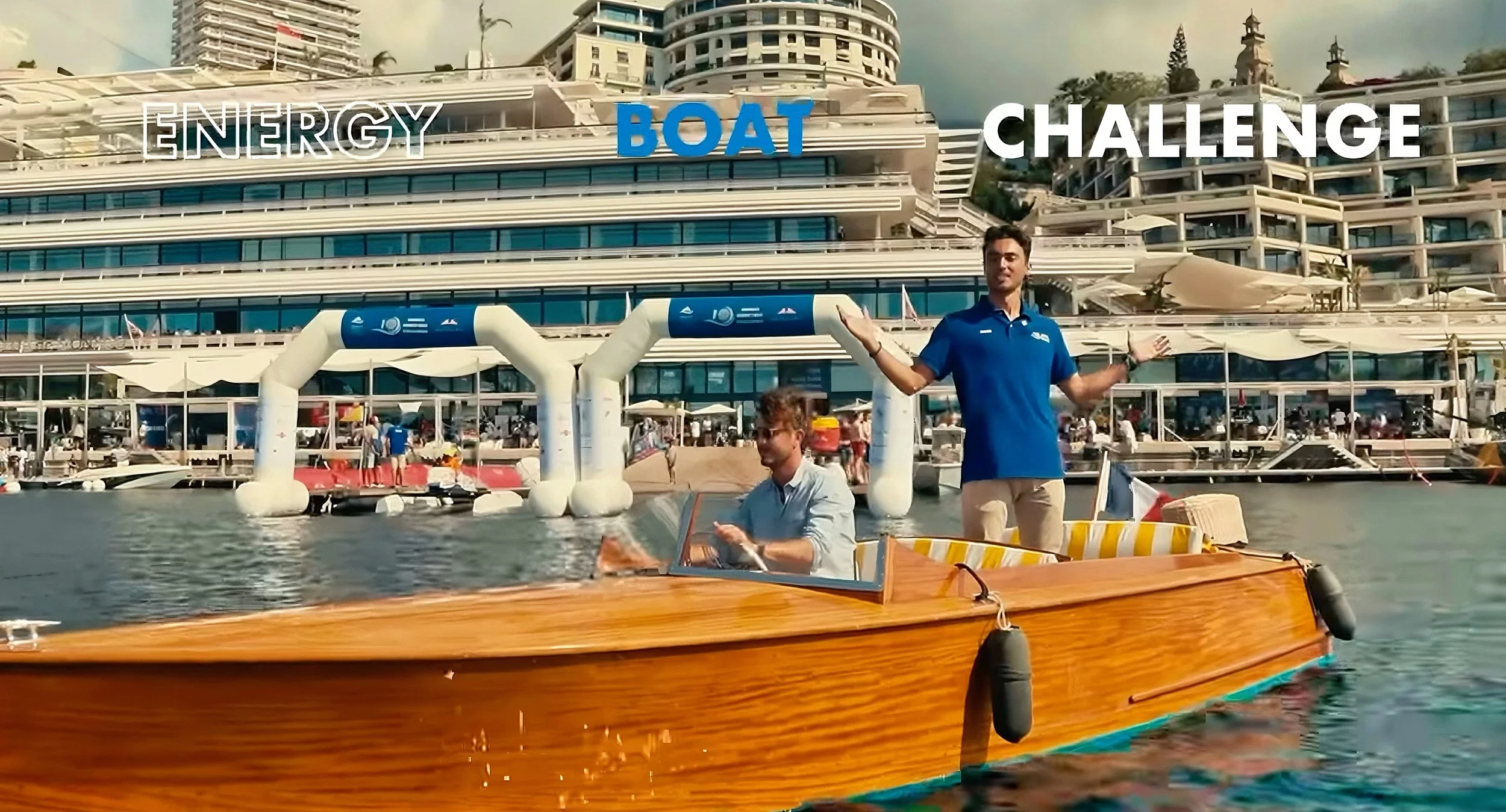 Two men on a wooden boat in a harbor, one sitting and one standing, with luxury yachts and a marina in the background. Overlay text reads 'Energy Boat Challenge'.