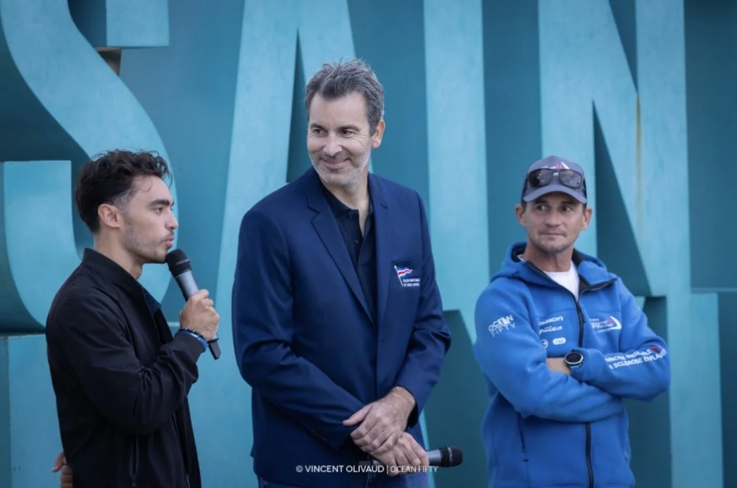 Three men standing outdoors in front of large teal-colored letters, one speaking into a microphone, the other two listening, with a light blue sky background.