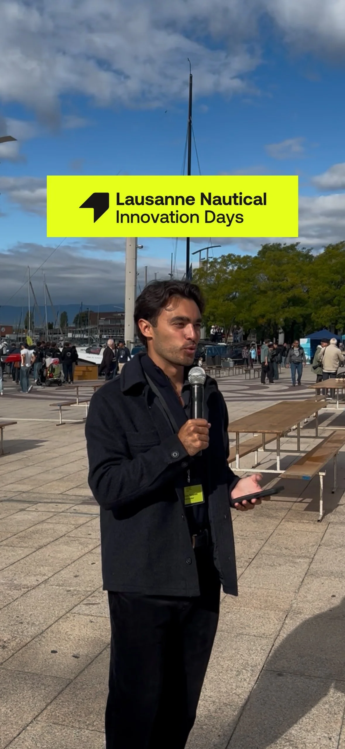 Nico Telenti holding a microphone and a phone, standing outdoors at Lausanne Nautical Innovation Days event, with people and boats in the background, under cloudy sky.